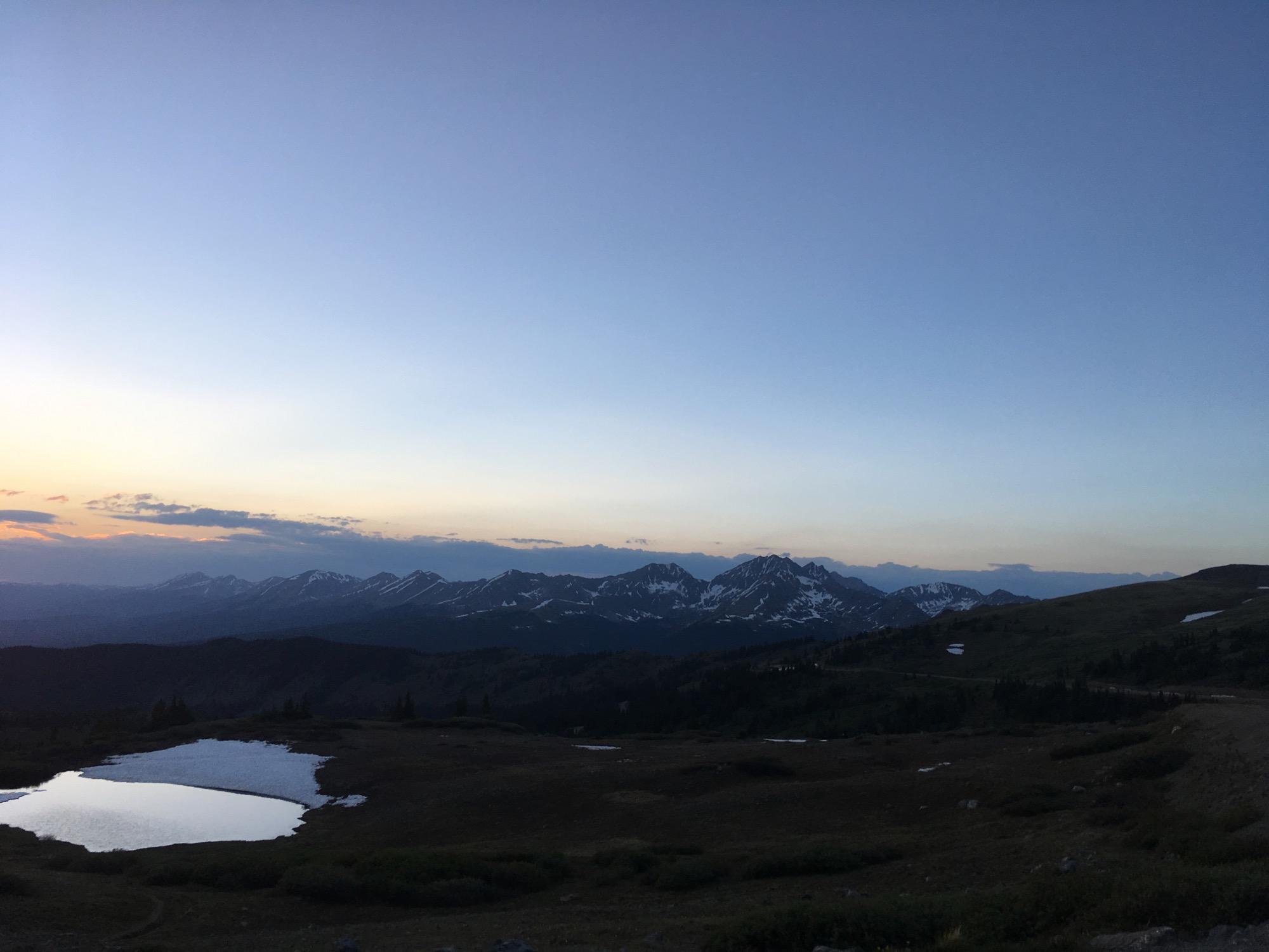A tranquil mountain landscape at dusk, featuring a calm body of water in the foreground, surrounded by lush greenery and rocky terrain. The sky transitions from soft blue to warm hues of orange and pink near the horizon, while snow-capped peaks rise in the distance, silhouetted against the fading light. Cottonwood Pass Road mountain bike trail.