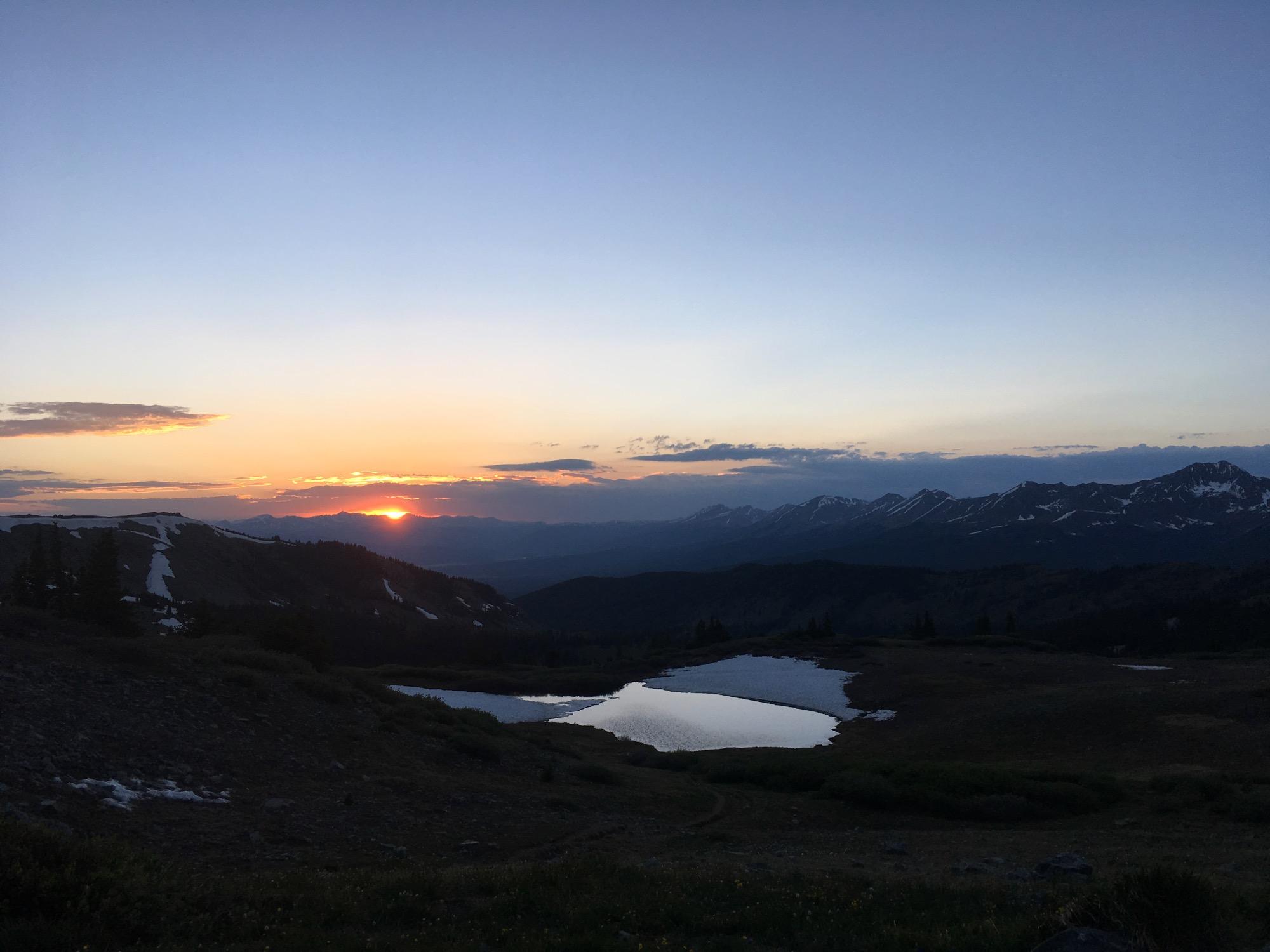 A serene mountain landscape at sunset, featuring a vibrant orange and pink sky. The sun is setting behind distant peaks, while a still, reflective body of water is visible in the foreground. Snow-capped mountains and rugged terrain add to the picturesque scene, with patches of green vegetation contrasting against the rocky ground. Cottonwood Pass Road mountain bike trail.