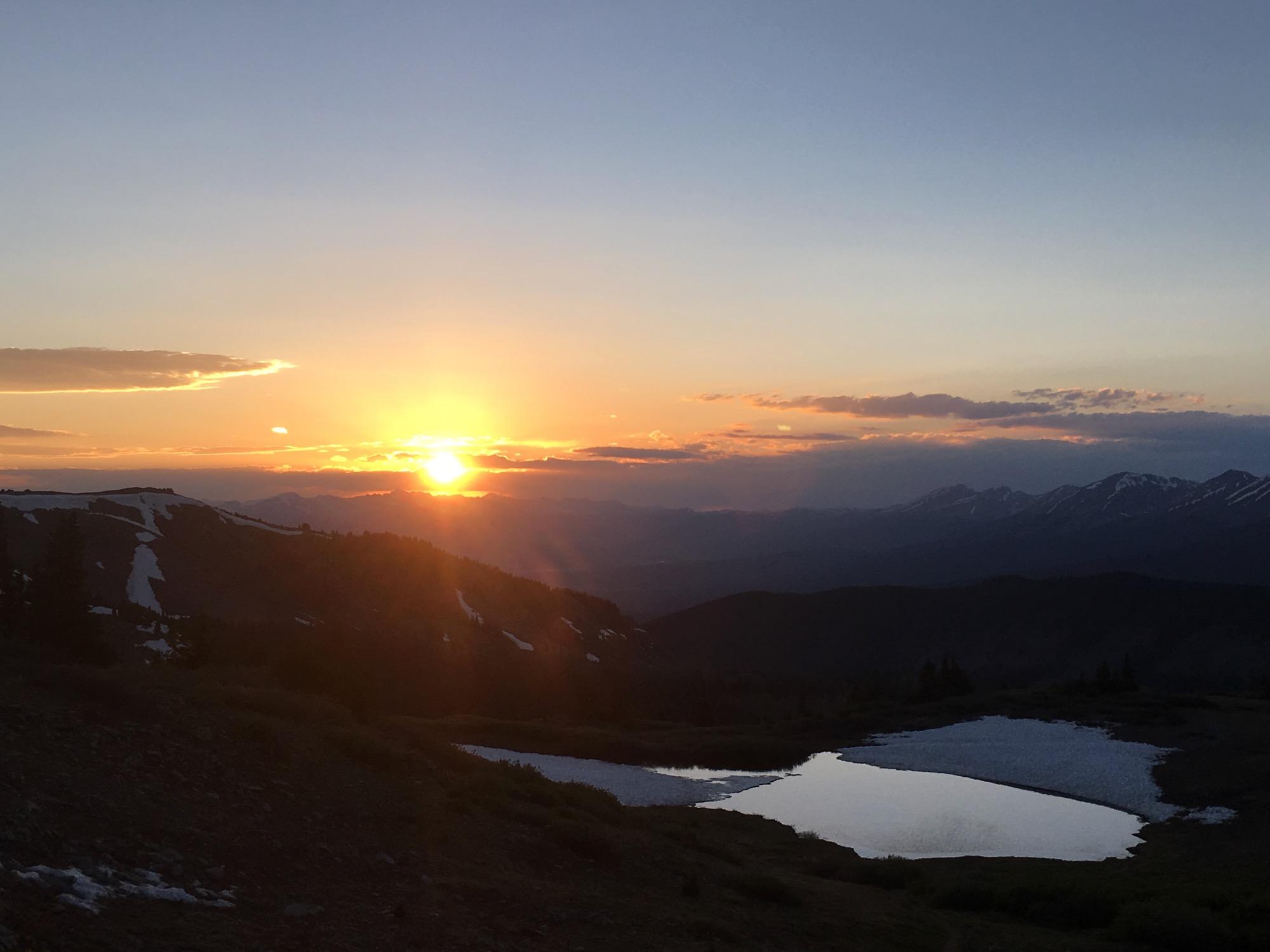 A breathtaking sunset over a mountainous landscape, featuring a vibrant orange sun dipping below the horizon. Silhouetted peaks and rolling hills create a dramatic backdrop, while a small reflective pond captures the golden light, enhancing the serene beauty of the scene. Additionally, patches of snow can be seen on the mountainsides, adding contrast to the warm colors of the sky. Cottonwood Pass Road mountain bike trail.