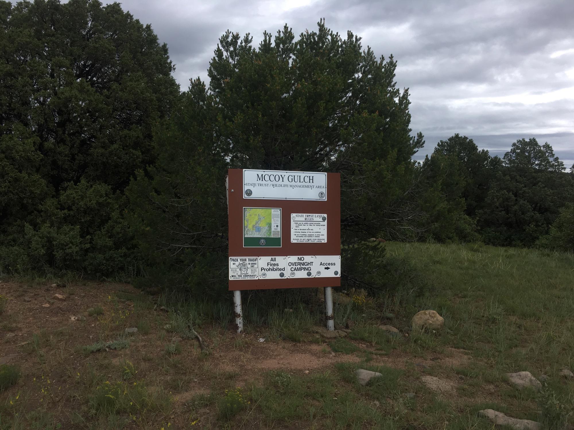 Signage for McCoy Gulch, a state trust and wildlife management area, featuring a map and regulations. Surrounded by greenery and cloudy skies. McCoy Gulch Road mountain bike trail.