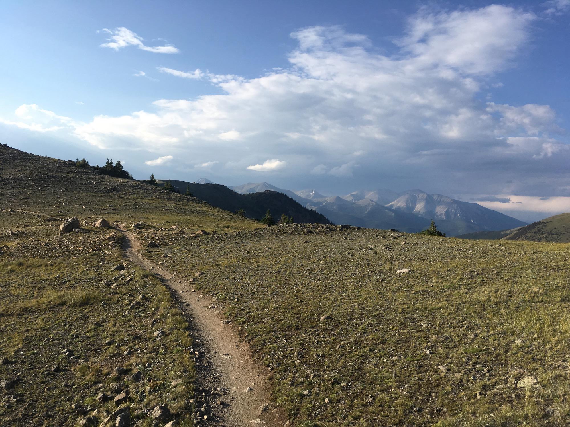 A winding dirt path leads through a rocky, grassy landscape with scattered boulders, surrounded by distant mountain peaks under a partly cloudy sky. Monarch Crest Trail mountain bike trail.