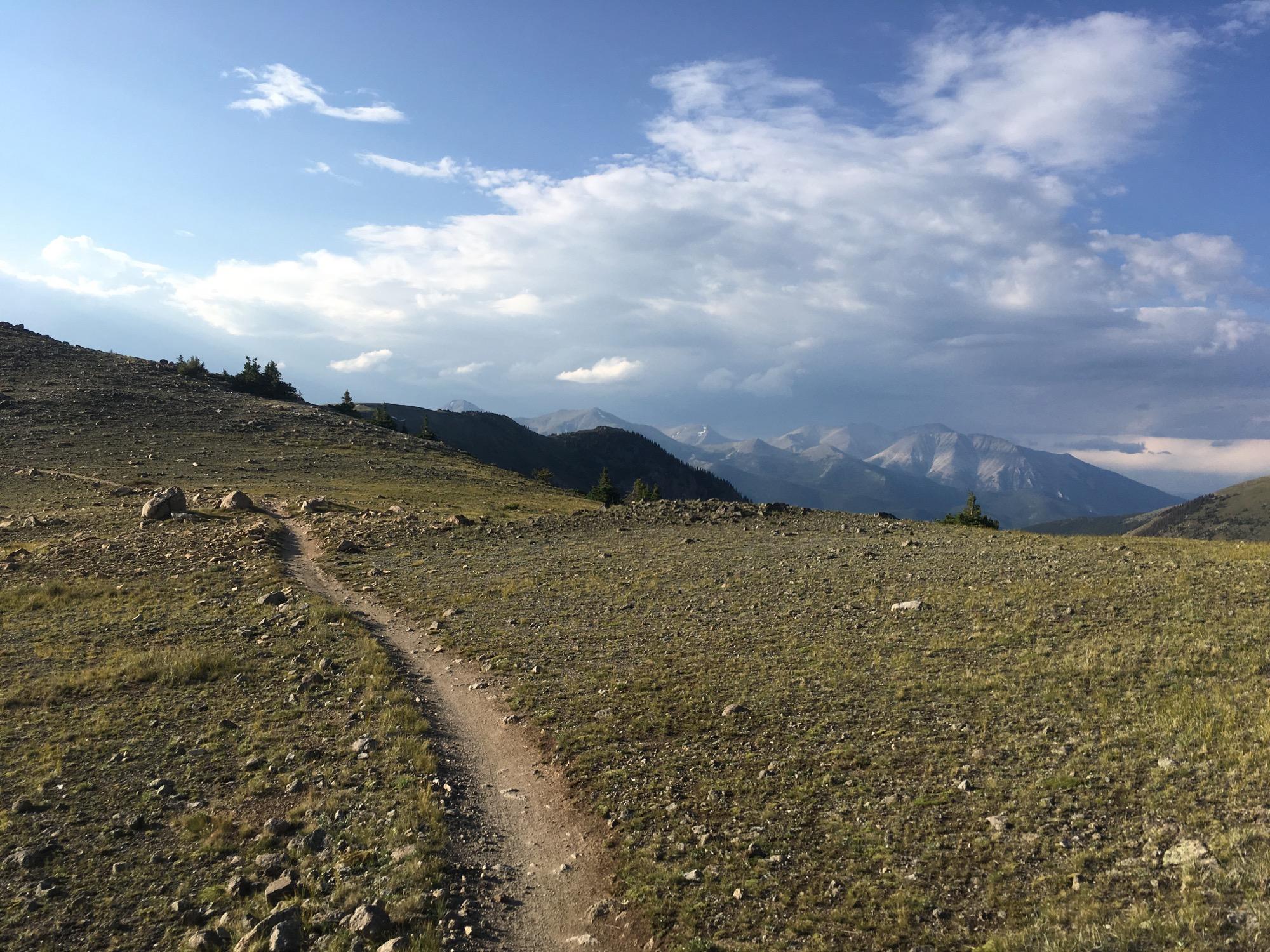 A winding dirt path leads through a rocky landscape, surrounded by grass and sparse trees, with distant mountains visible under a partly cloudy sky. Monarch Crest Trail mountain bike trail.