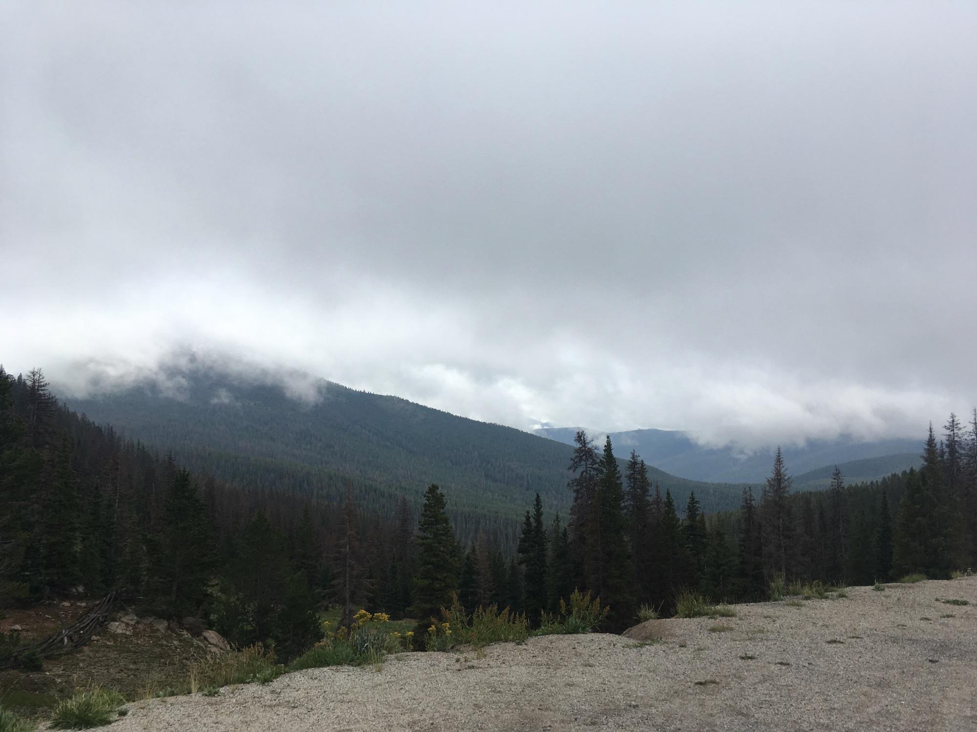 A cloudy landscape view of mountains covered in evergreen trees, with mist hovering over the peaks and a gravel foreground. The sky is overcast, indicating a potentially rainy day in a natural outdoor setting. Monarch Crest Trail mountain bike trail.