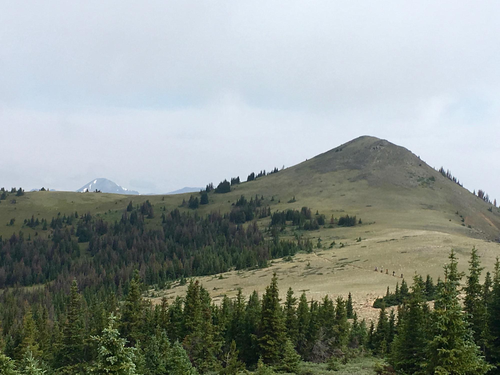 A view of rolling green hills with a prominent peak in the background. The landscape features patches of trees and a few hikers ascending the slope, with a cloudy sky overhead. Snow-capped mountains are faintly visible in the distance. Monarch Crest Trail mountain bike trail.