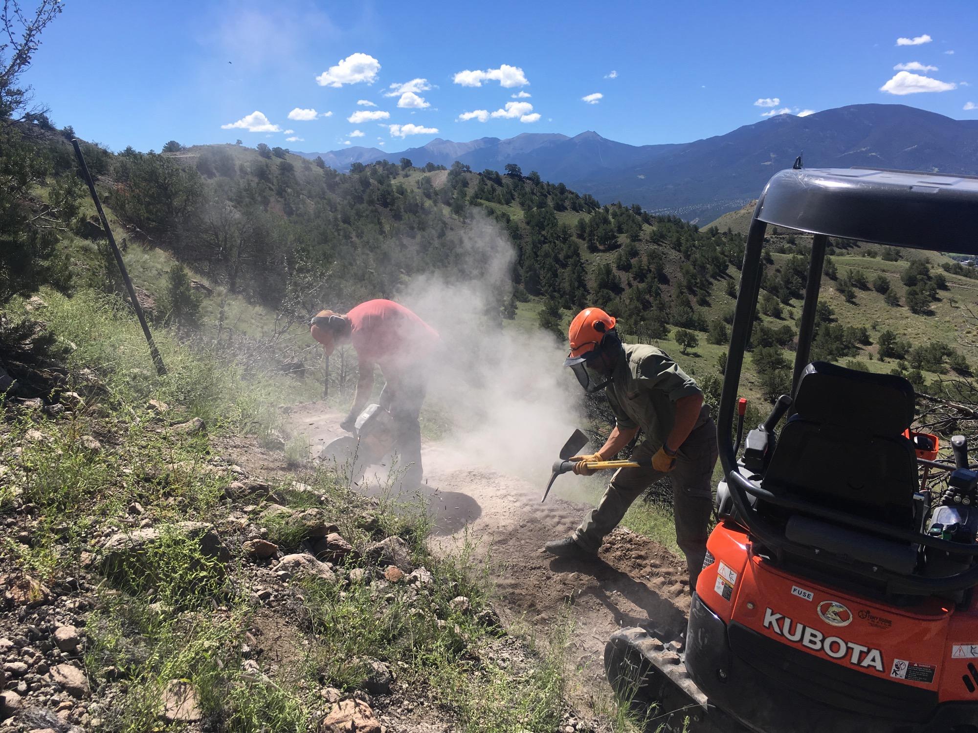 Two workers are using tools on a rugged landscape, creating dust and debris. One is bent over, working with a circular saw, while the other stands beside him, using a shovel. A small orange Kubota vehicle is parked nearby. The scene is set against rolling hills and a blue sky with scattered clouds. Trees and vegetation surround the area, indicating a natural outdoor environment. Rusty Lung mountain bike trail.