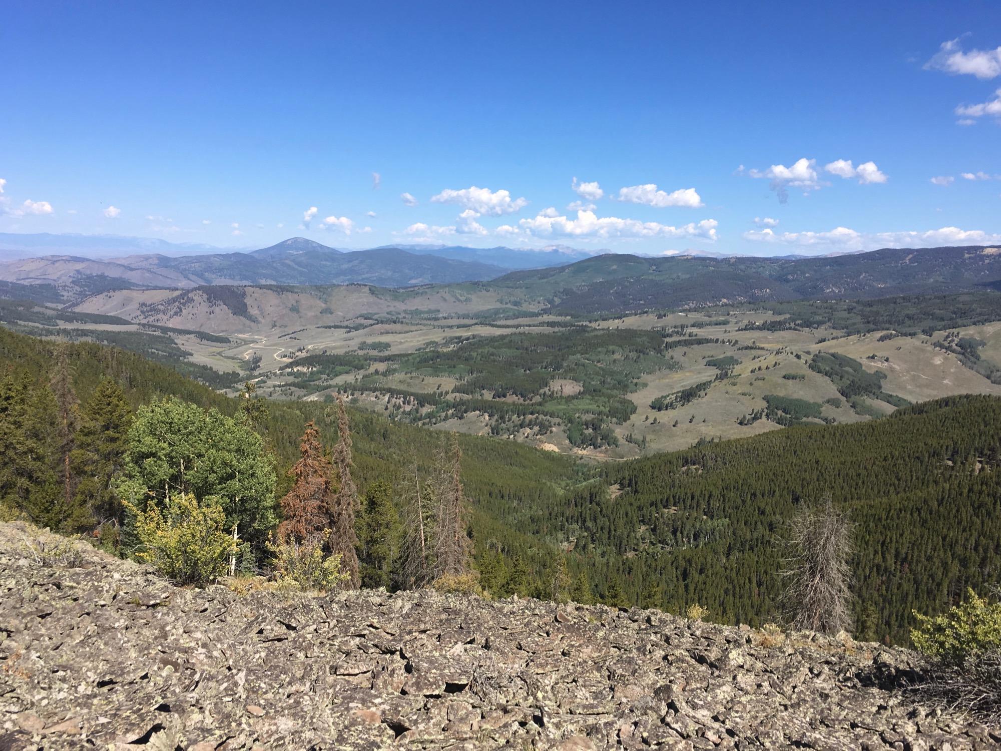 A panoramic view of a mountainous landscape under a clear blue sky, featuring rolling green hills, scattered trees, and a winding road in the distance. The foreground includes rocky terrain with various rock formations, while the background showcases multiple layers of mountains and patches of clouds. Colorado Trail: Marshall Pass to Sargents Mesa / Rd #855 mountain bike trail.