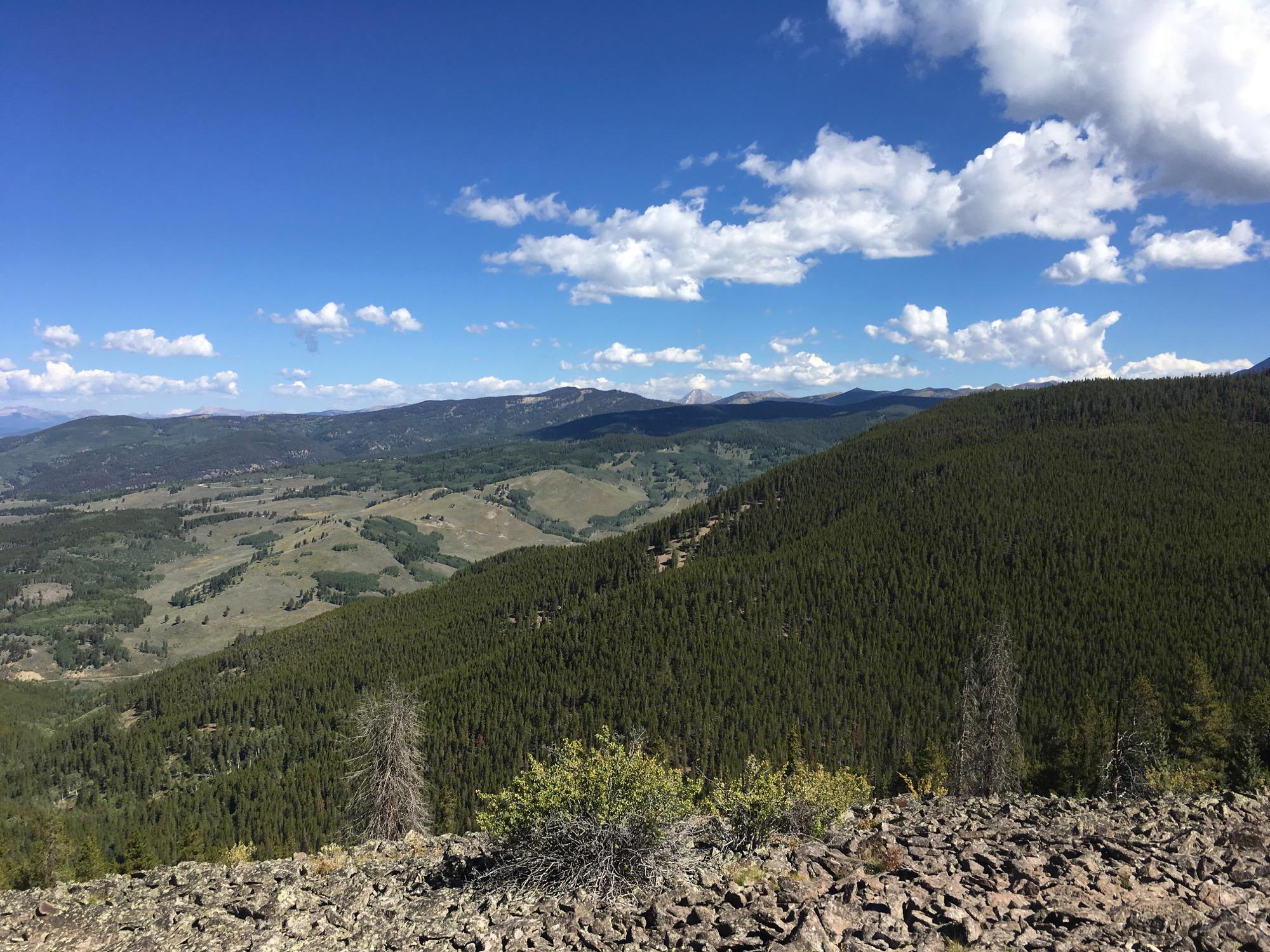 A panoramic view of a mountainous landscape featuring rolling hills covered in lush greenery, interspersed with patches of rocky terrain. The sky is bright blue with scattered fluffy white clouds, creating a serene and tranquil atmosphere. The distant mountains are visible under the clear sky, showcasing a beautiful natural environment. Colorado Trail: Marshall Pass to Sargents Mesa / Rd #855 mountain bike trail.