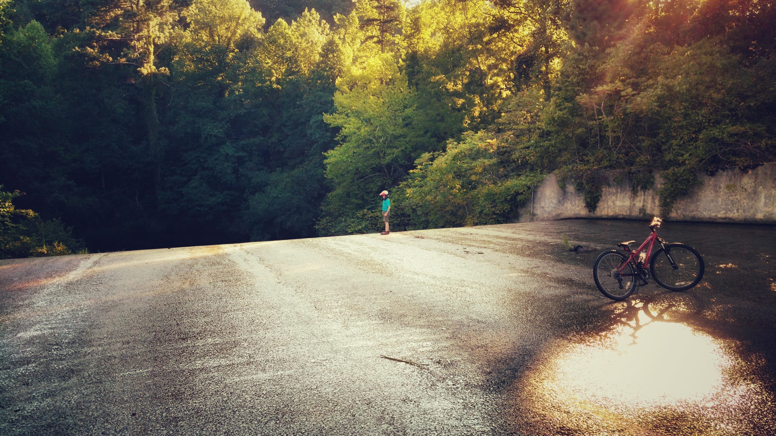 A scenic view of a person standing on a sunlit, wet surface surrounded by lush green trees. A bicycle rests nearby in the foreground, reflecting the warm light of the setting sun. The scene conveys a sense of tranquility and connection with nature. Lake Russell Loop mountain bike trail.