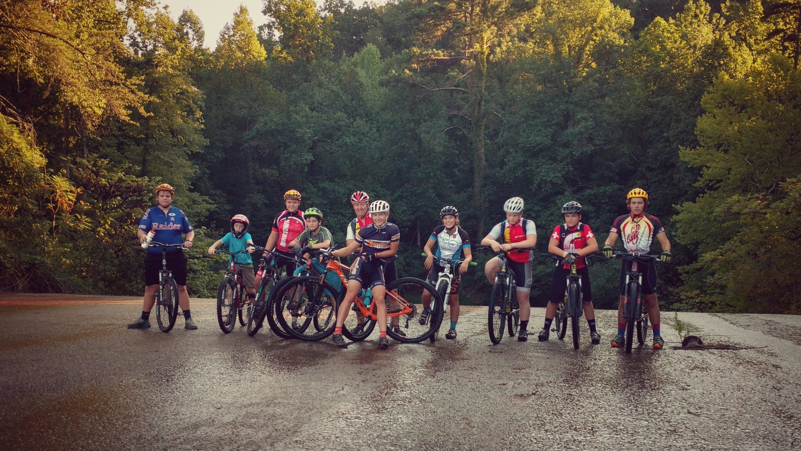 A group of young cyclists poses on a wet road surrounded by lush green trees. They are wearing colorful cycling jerseys and helmets, with each person having their own bicycle. The scene captures a moment of camaraderie and enthusiasm for biking outdoors. Lake Russell Loop mountain bike trail.