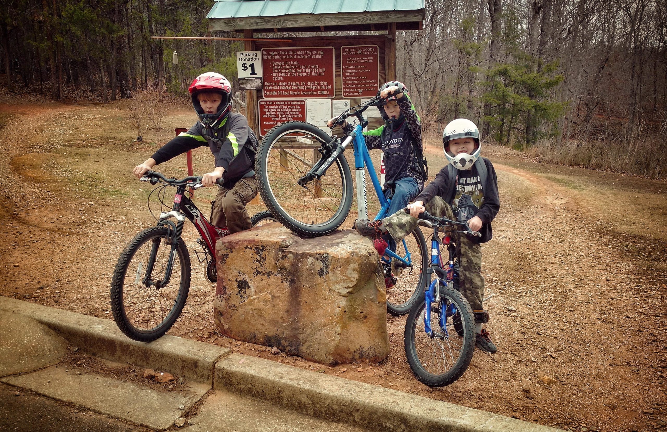 Three kids in helmets pose with their bicycles on a rock near a trailhead sign in a wooded area. The boy on the left is holding a red mountain bike, while the other two are on their bikes, one blue and one smaller blue. The sign behind them indicates a parking donation fee and provides information about the surrounding trails. Chicopee Woods mountain bike trail.