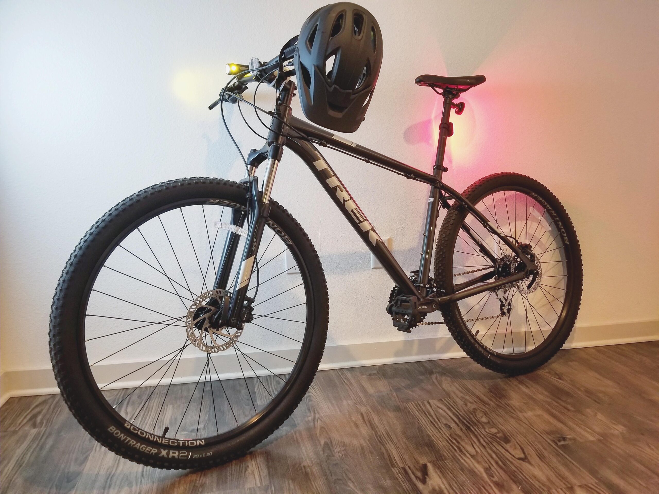 Trek Marin 6: A black Trek mountain bike is leaning against a white wall, featuring a helmet attached to the handlebars. The bike is equipped with front headlights and red rear lights, highlighting its safety features. The setting includes a wooden floor that complements the bike's design.