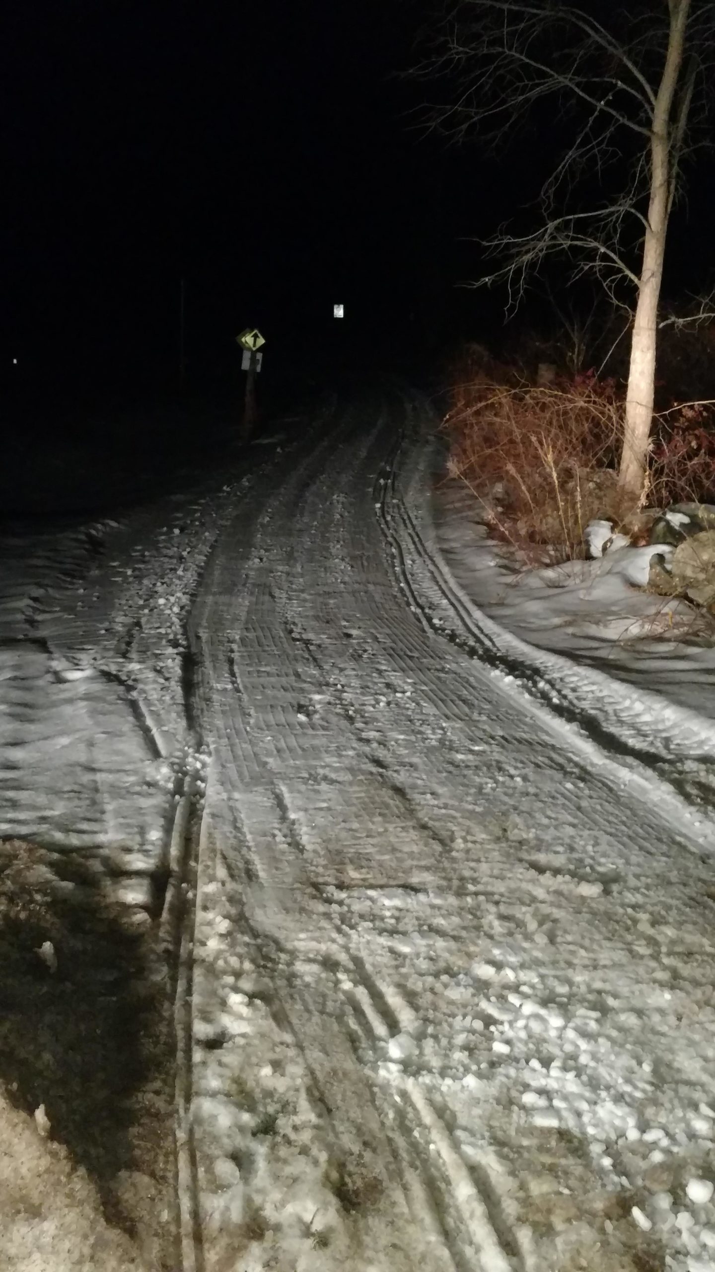A dirt road covered in snow and ice, illuminated by minimal lighting at night, with a fork in the road and a sign visible in the background. Sparse vegetation and trees line the sides of the road. Lake Auburn Loop Trail mountain bike trail.