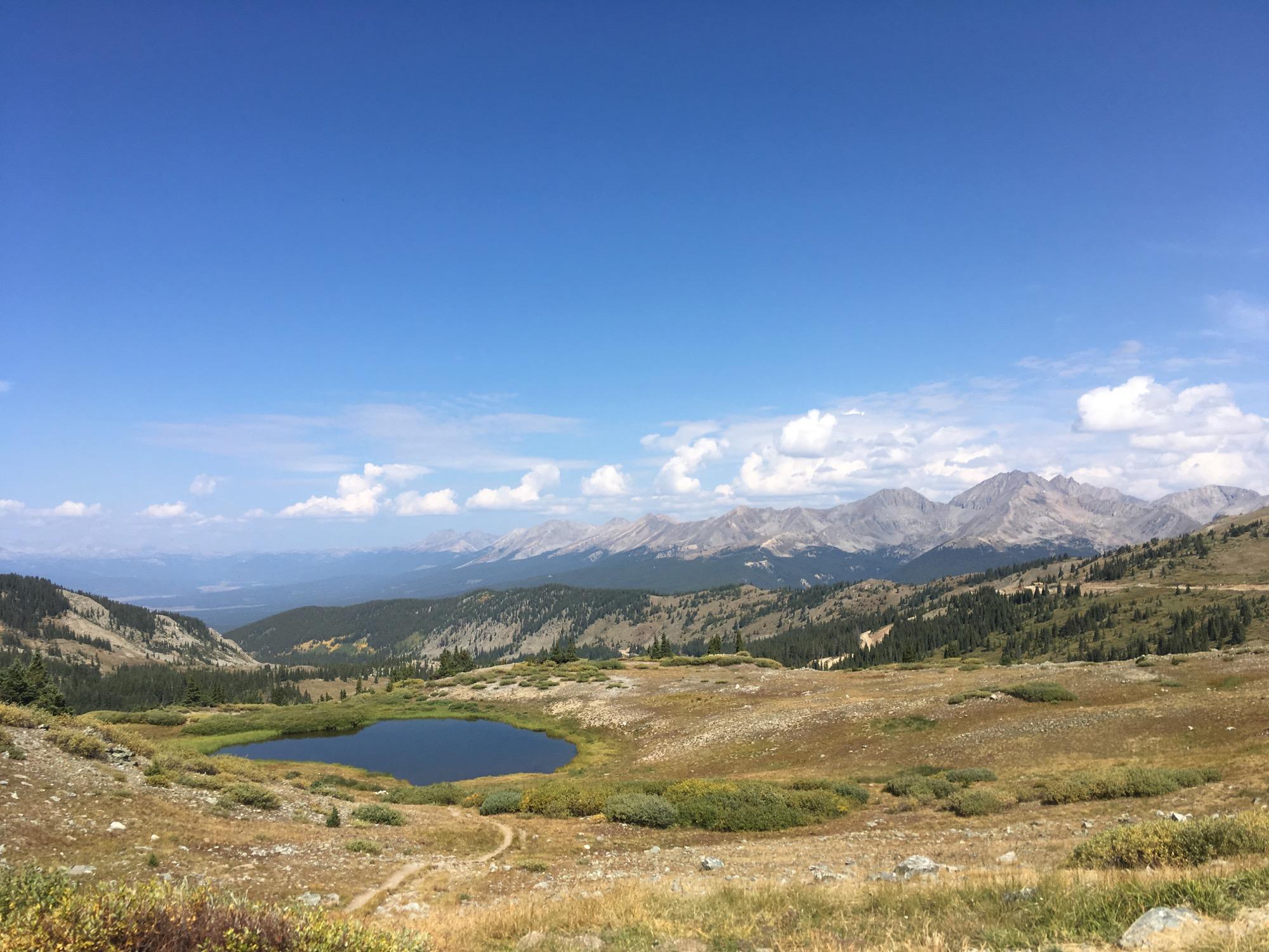 A panoramic view of a mountainous landscape featuring rolling hills, a small tranquil pond, and a bright blue sky with fluffy white clouds. Pine trees dot the slopes, and the rugged mountain peaks rise majestically in the background. Cottonwood Pass Road mountain bike trail.