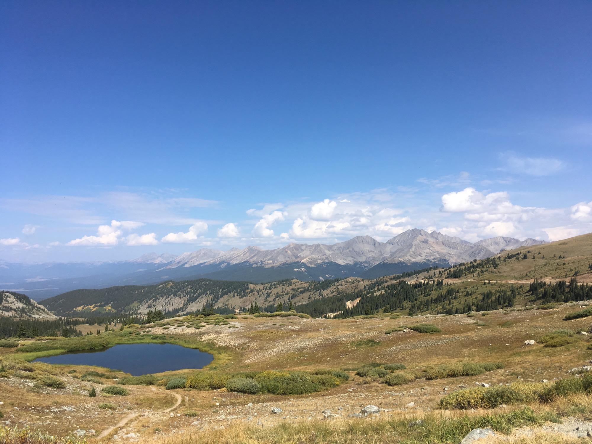 A scenic view of a mountain landscape featuring rolling hills, a clear blue sky with fluffy white clouds, and a tranquil pond surrounded by greenery. In the background, majestic mountains rise dramatically against the horizon, creating a picturesque natural setting. Cottonwood Pass Road mountain bike trail.