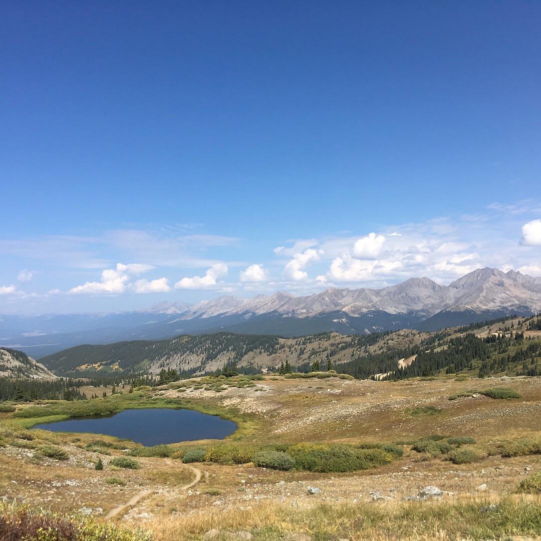 A scenic view of a mountainous landscape featuring a tranquil pond surrounded by grassy fields and shrubs, under a bright blue sky scattered with fluffy white clouds. The distant mountains create a stunning backdrop, showcasing varying shades of green and brown in the foreground and rugged peaks in the background. Cottonwood Pass Road mountain bike trail.