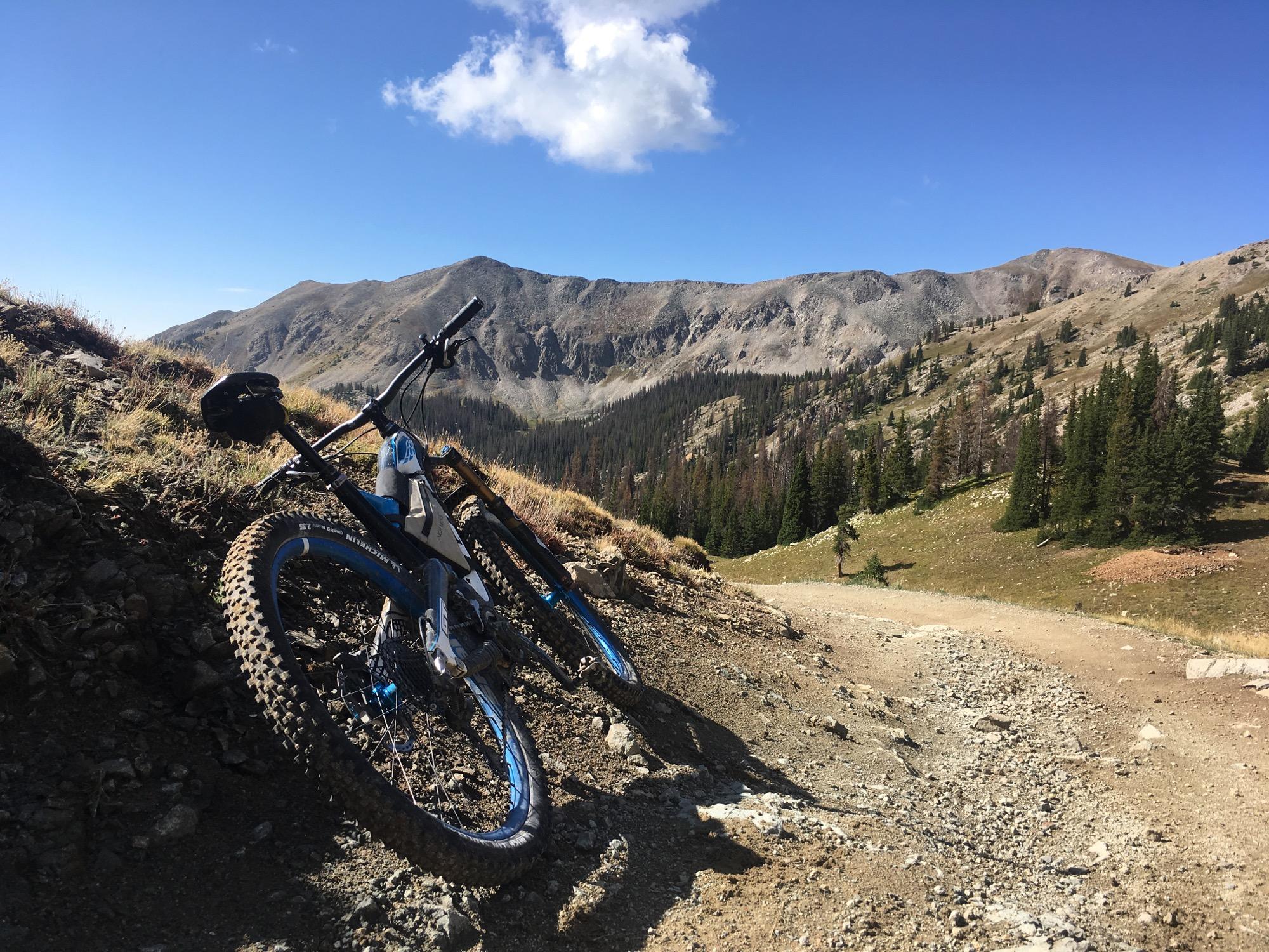 Two mountain bikes are leaning against a rocky incline on a dirt trail, with a backdrop of rolling hills and a clear blue sky. Pine trees dot the landscape, and the mountains in the distance add to the scenic outdoor setting. Canyon Creek Trail mountain bike trail.