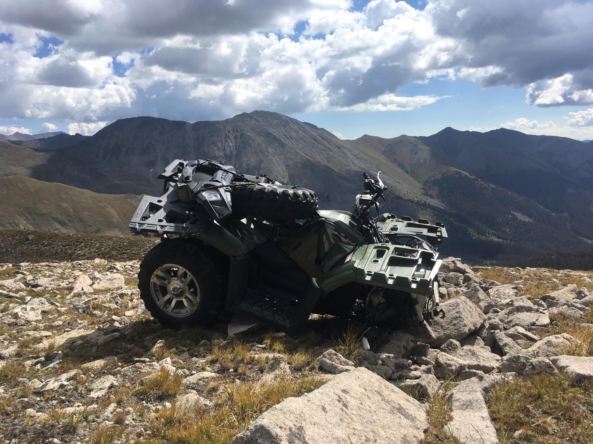 An all-terrain vehicle (ATV) is parked on rocky terrain with mountainous hills in the background under a partly cloudy sky. The landscape is rugged, featuring sparse vegetation and a distant view of additional peaks. Canyon Creek Trail mountain bike trail.