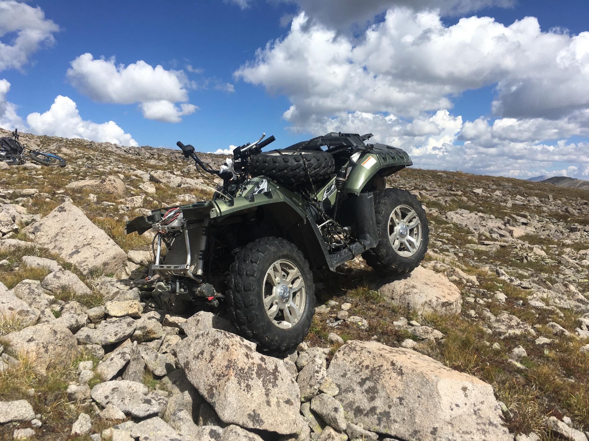 An all-terrain vehicle (ATV) parked on rocky terrain with a cloudy blue sky in the background. The ATV is green with large, rugged tires, and the surrounding landscape features scattered rocks and grass. Canyon Creek Trail mountain bike trail.