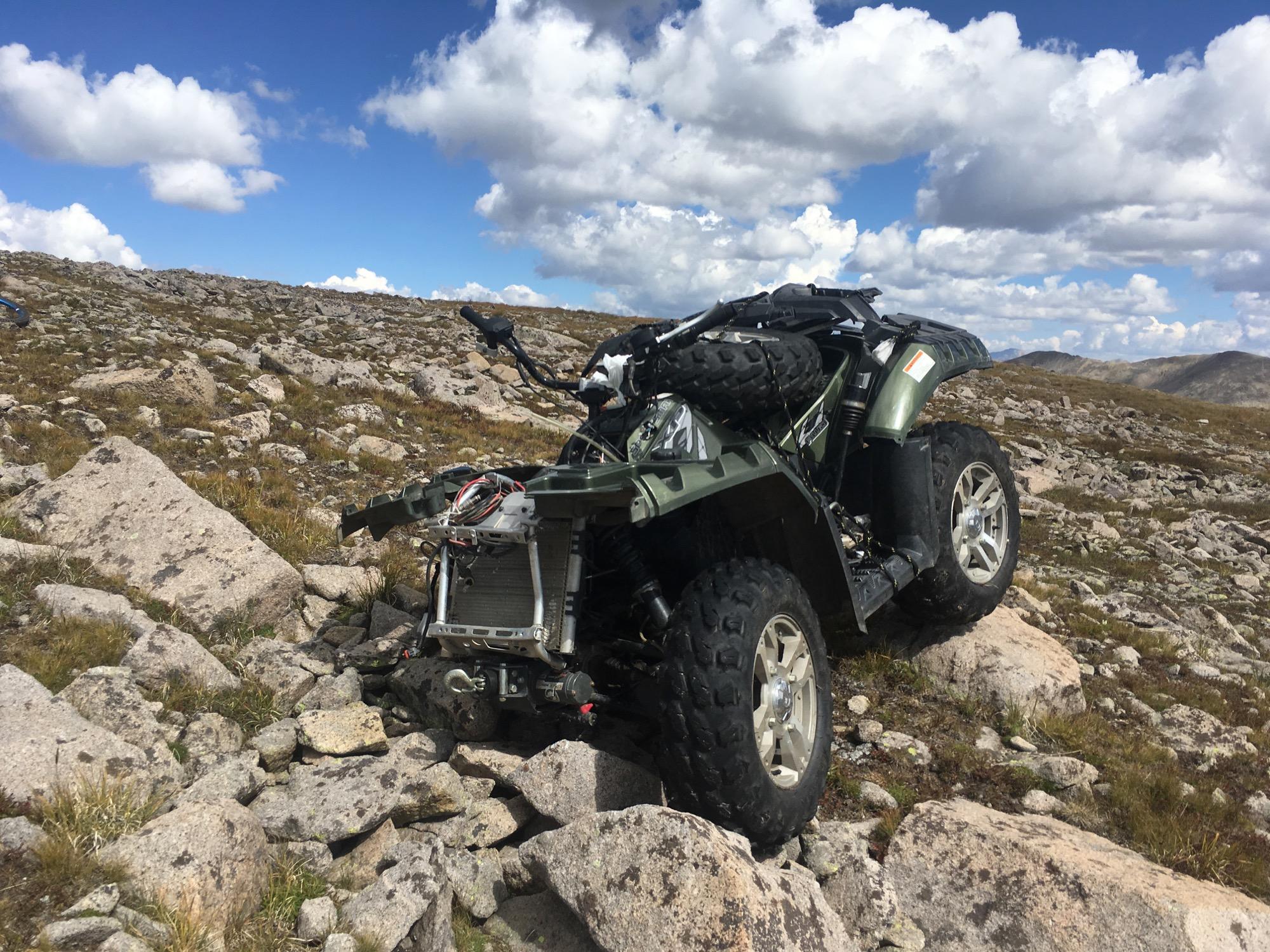 An all-terrain vehicle (ATV) rests on rocky terrain under a partly cloudy sky. The vehicle is positioned at an angle, showcasing its rugged tires and green frame. Surrounding it are large boulders and patches of grass, with distant mountain ranges visible in the background. Canyon Creek Trail mountain bike trail.