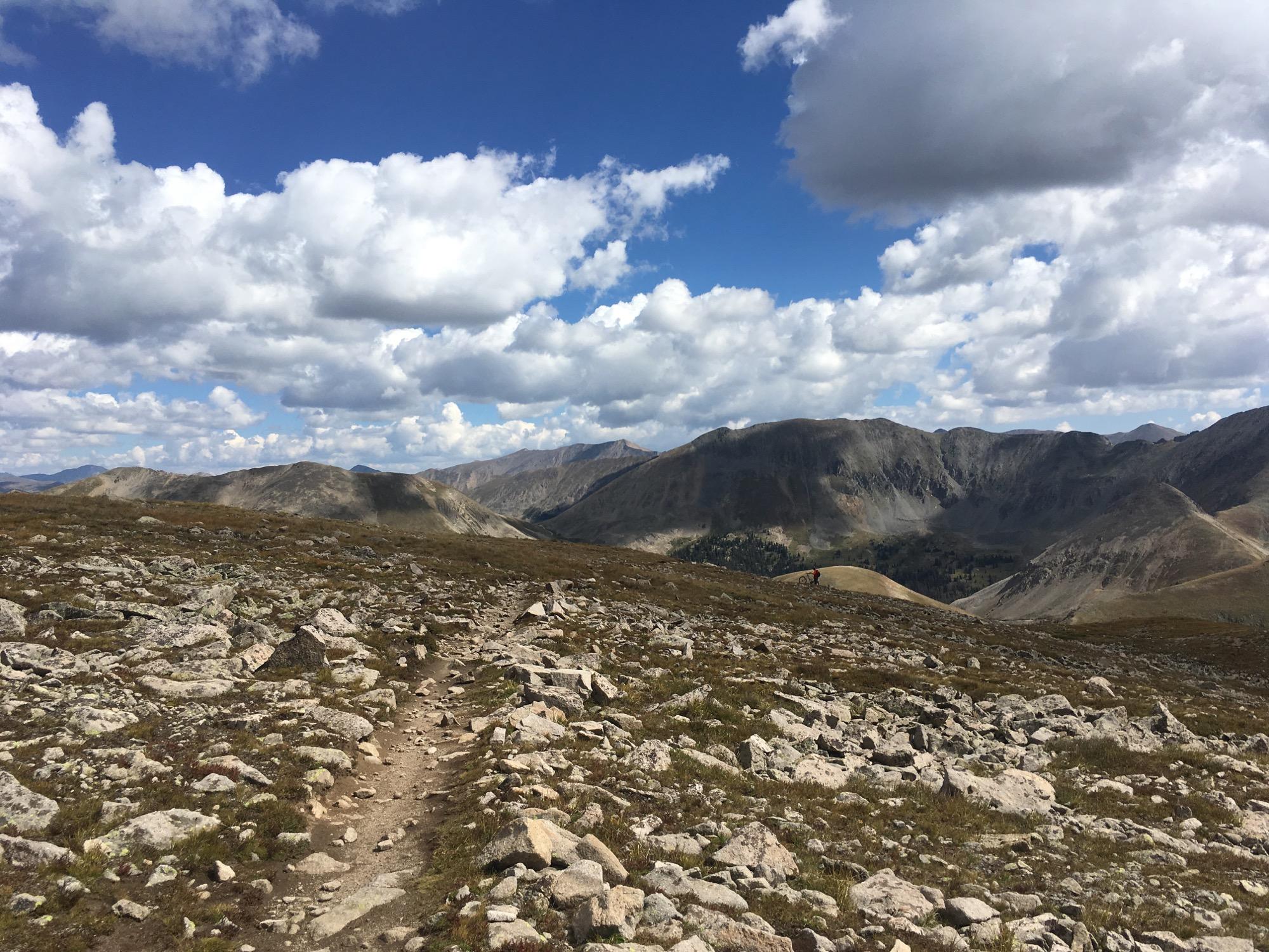 A rocky trail meanders through a vast mountainous landscape under a blue sky filled with white clouds. The scene features rolling hills and distant mountains, showcasing a mix of grassy and rocky terrain typical of high-altitude environments. Canyon Creek Trail mountain bike trail.