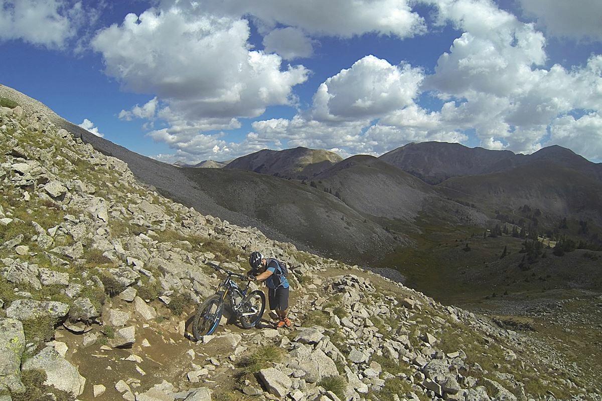 A mountain biker troubleshooting their bike on a rocky trail in a mountainous landscape, with hills and blue sky adorned with clouds in the background. Canyon Creek Trail mountain bike trail.