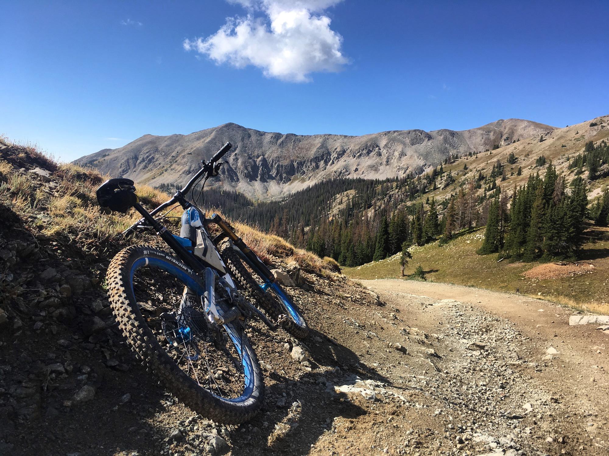 Two mountain bikes lean against a rocky terrain, with a backdrop of rugged mountains and a clear blue sky. The surrounding landscape features a mix of grassy areas and evergreen trees, indicating a high-altitude environment. Canyon Creek Trail mountain bike trail.