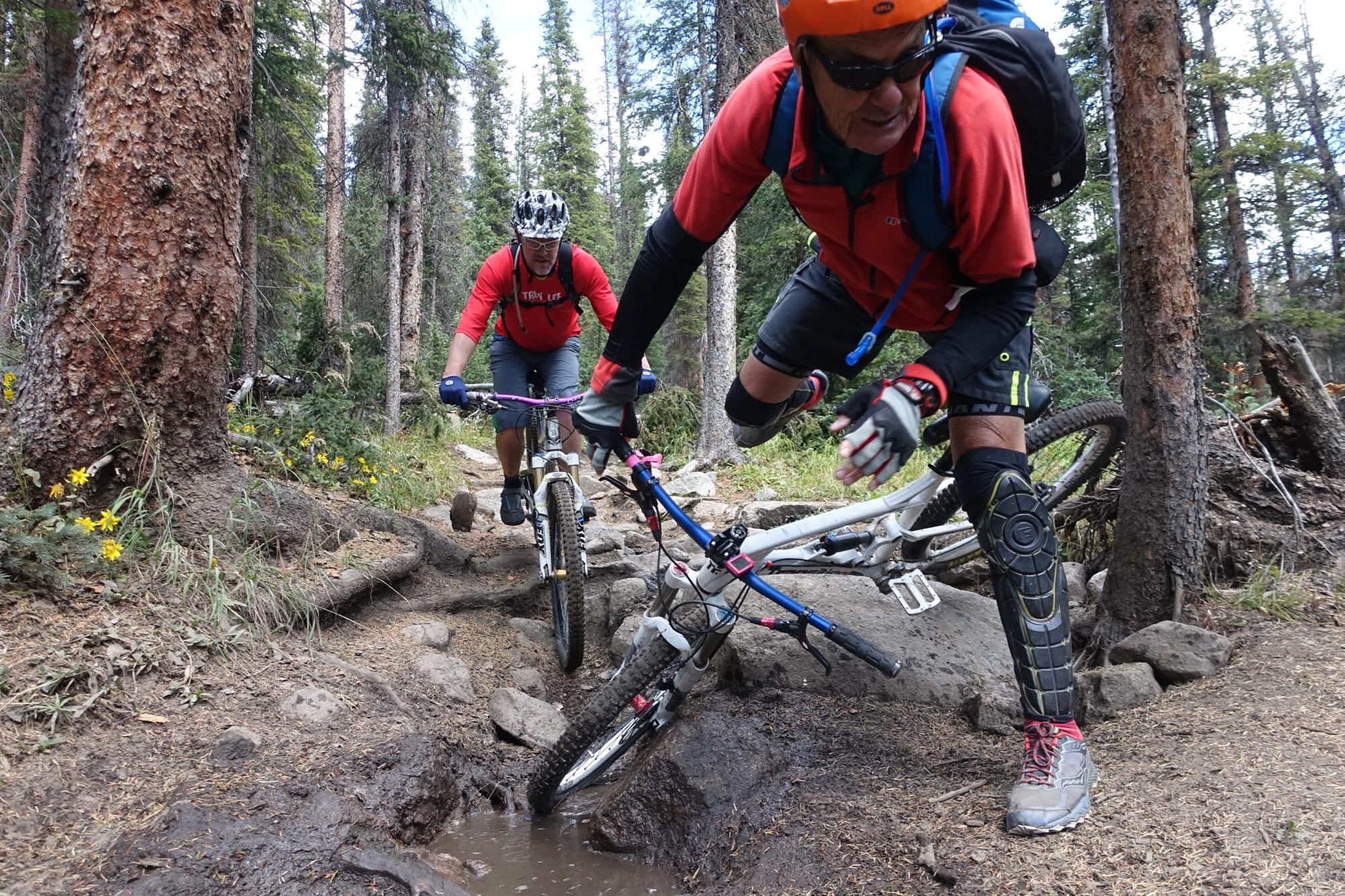 Two mountain bikers navigating a rocky trail in a forested area. One rider is airborne, leaping over a muddy patch, while the other follows closely behind. Both are wearing helmets and protective gear, with the surrounding landscape featuring tall trees and wildflowers. Canyon Creek Trail mountain bike trail.