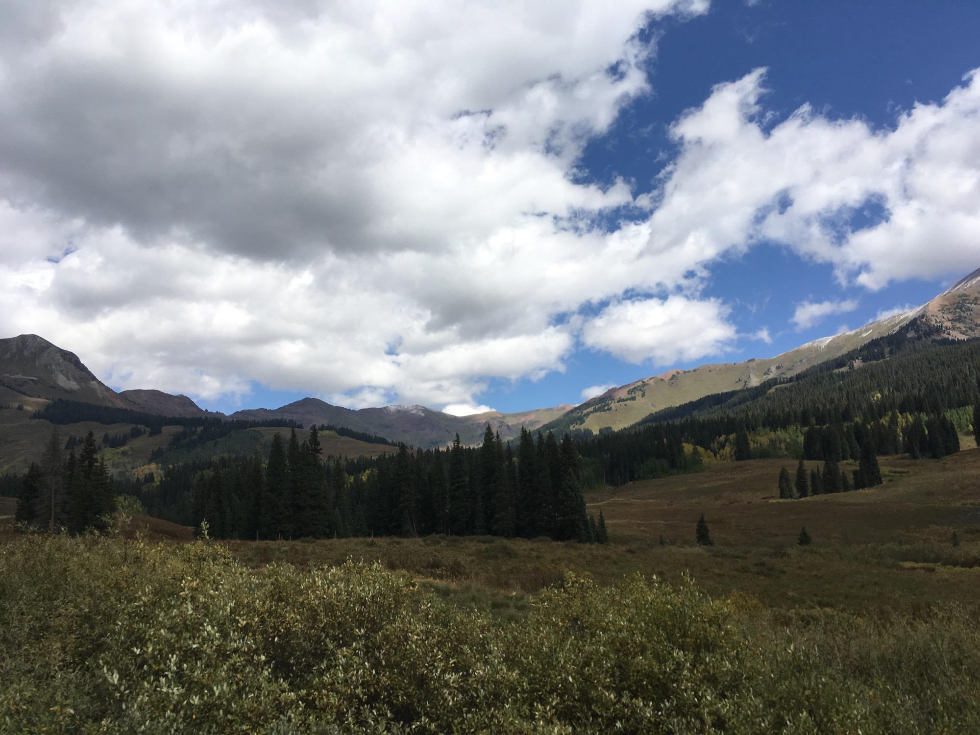 A scenic landscape featuring rolling mountains under a partly cloudy sky, with patches of blue peeking through. Dense evergreen trees cover the foreground, and vibrant fall colors can be seen on the mountainsides in the background. Washington Gulch Road mountain bike trail.