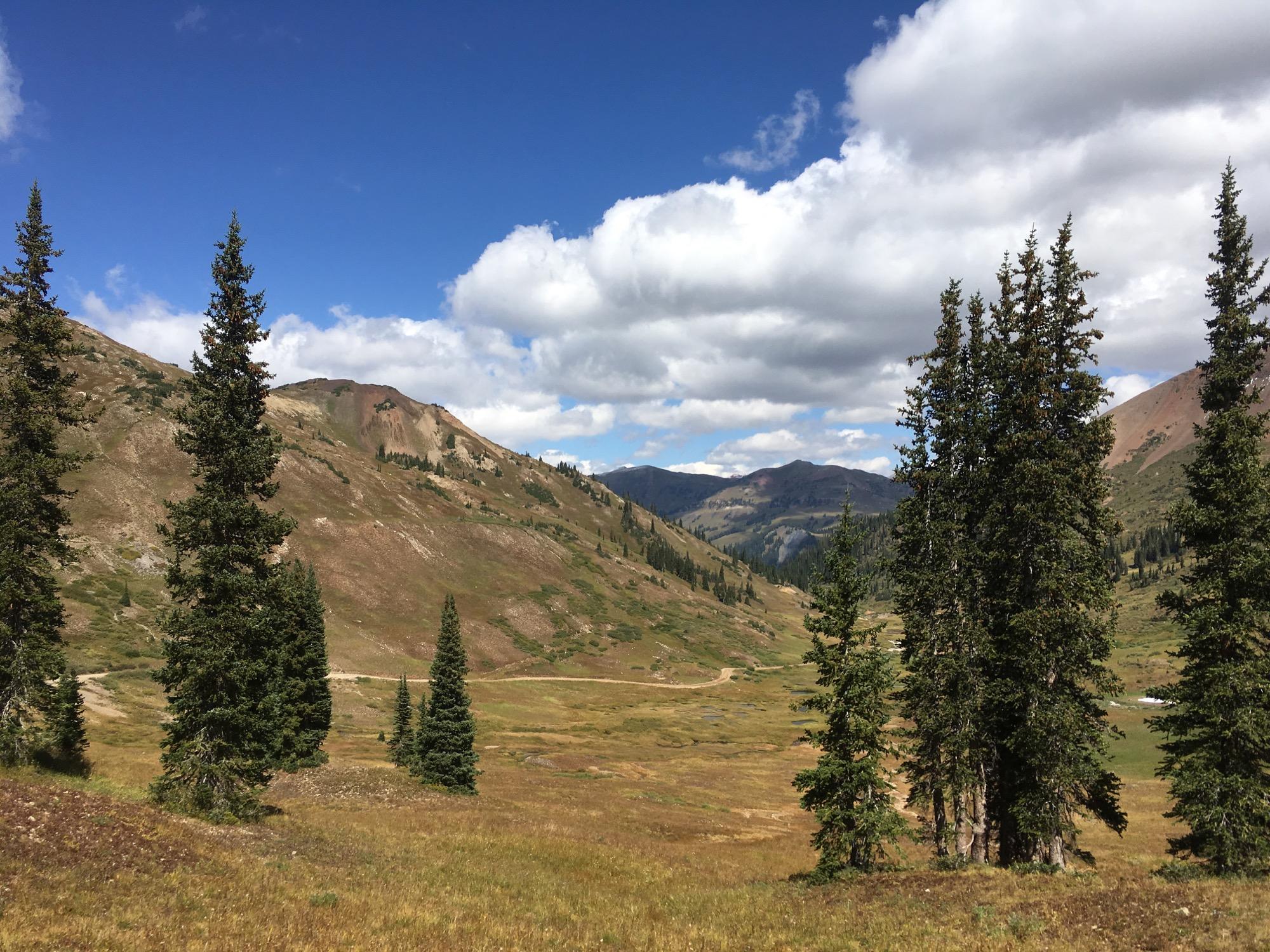A picturesque mountain landscape featuring tall evergreen trees in the foreground, with rolling hills and vibrant greenery in the background. The sky is bright blue, dotted with fluffy white clouds. A winding dirt road can be seen meandering through the valley, flanked by rocky hills and distant mountain peaks. Slate River Road mountain bike trail.