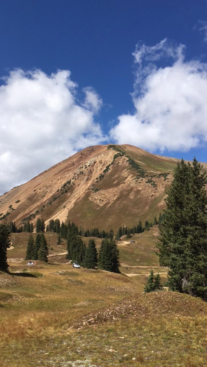 A scenic view of a mountainous landscape under a bright blue sky, featuring a prominent ridge covered in patches of greenery. In the foreground, grassy hills lead up to a mix of coniferous trees, with a few vehicles visible along a winding dirt road. Fluffy white clouds float above the peak, adding to the serene natural beauty of the scene. Slate River Road mountain bike trail.