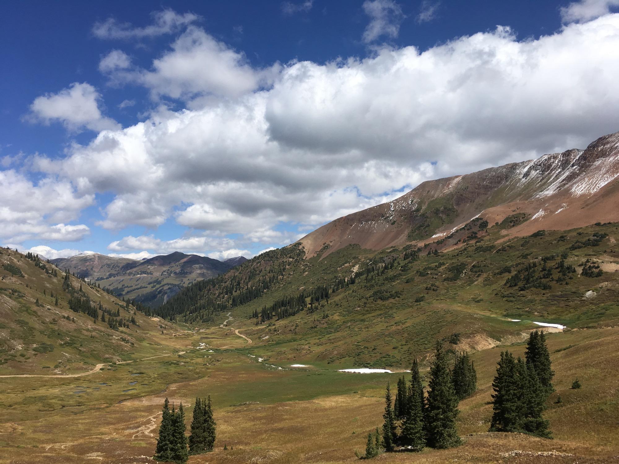 A breathtaking view of a mountainous landscape with rolling hills, lush green valleys, and patches of snow. The sky is bright blue with fluffy white clouds, highlighting the natural beauty of the region. Pine trees dot the foreground, and a winding dirt path leads through the greenery, inviting exploration. Slate River Road mountain bike trail.
