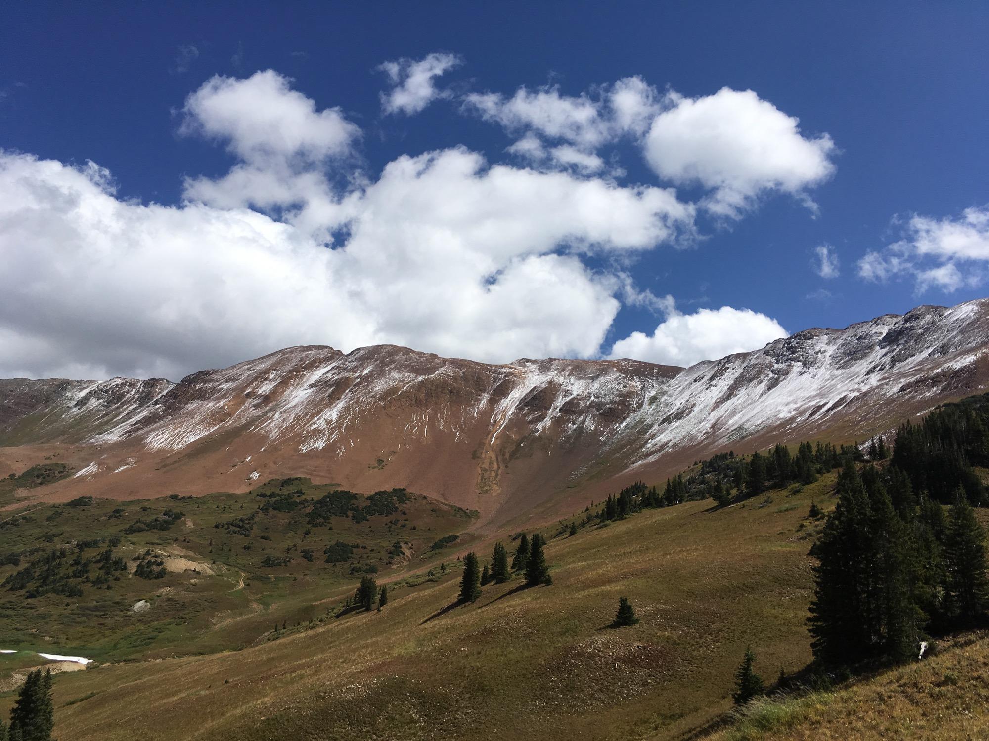 A picturesque mountain landscape featuring rugged, snow-dusted peaks under a bright blue sky with fluffy white clouds. The foreground showcases grassy slopes dotted with evergreen trees, while the mountains display a mix of brown and white terrain, indicating a transition from summer to early winter. Slate River Road mountain bike trail.