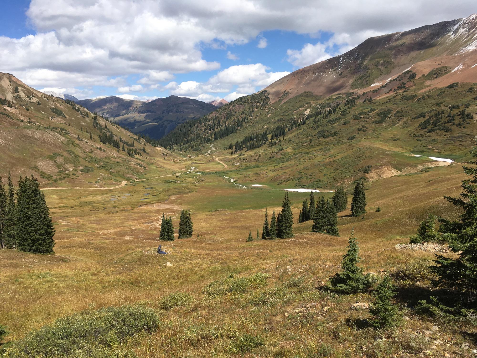 A panoramic view of a mountainous landscape featuring a wide valley with patches of green grass, scattered trees, and distant snowy peaks under a partly cloudy sky. Dirt paths meander through the valley, leading into the horizon. Slate River Road mountain bike trail.