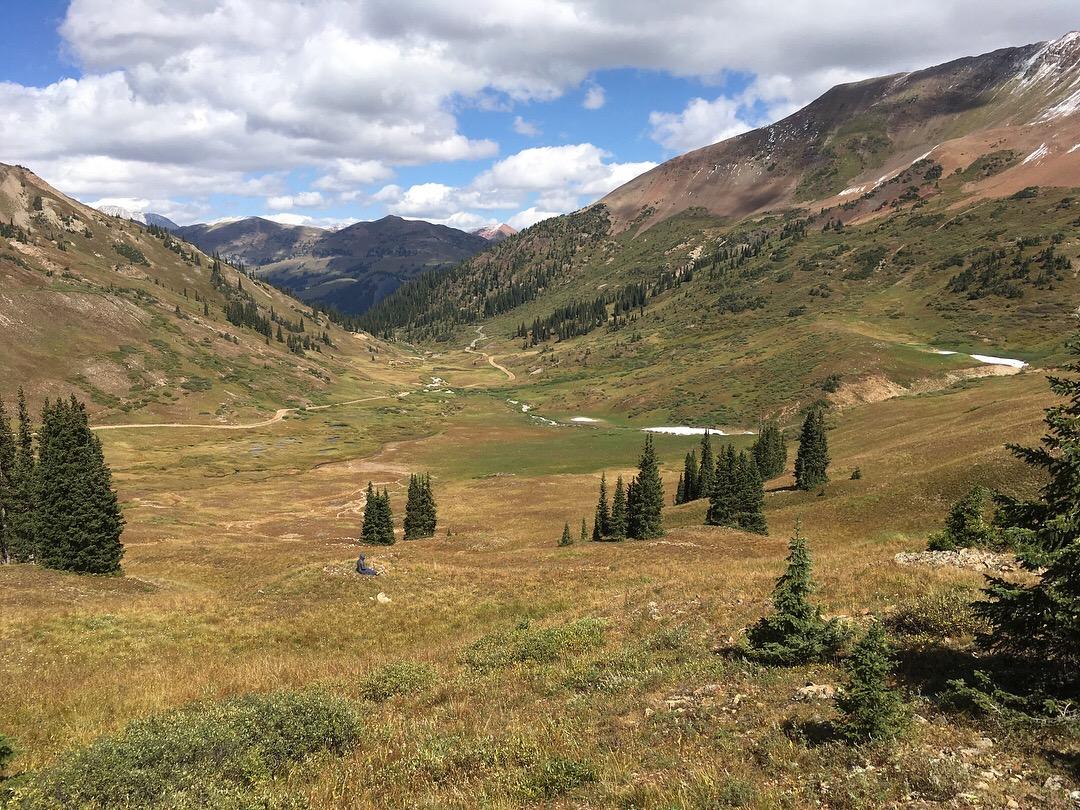 A scenic mountain valley landscape featuring lush green grass and patches of yellow wildflowers. Tall evergreen trees dot the foreground, while rolling hills and distant mountain peaks provide a dramatic backdrop under a partly cloudy sky. A winding path can be seen meandering through the valley. Slate River Road mountain bike trail.