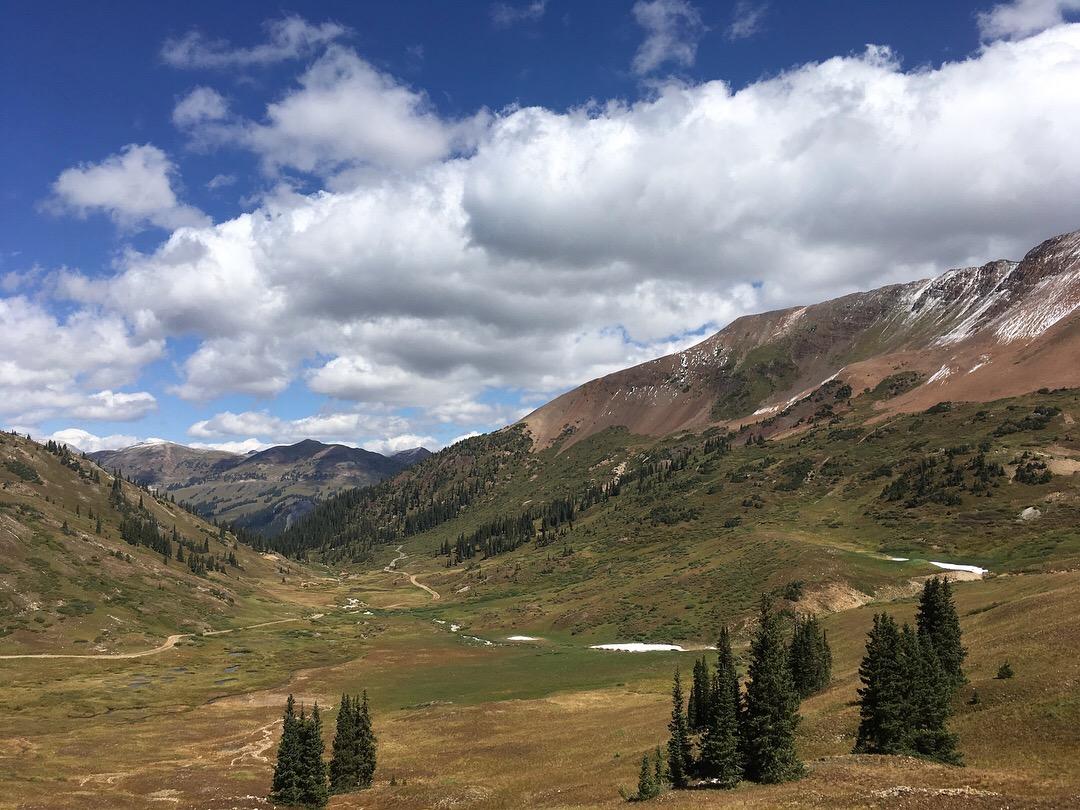 A panoramic view of a mountainous landscape featuring rolling hills, green valleys, and scattered patches of snow. The scene is under a bright blue sky dotted with fluffy white clouds, with pine trees situated in the foreground and rugged mountain peaks in the background. A winding dirt path can be seen cutting through the valley. Slate River Road mountain bike trail.