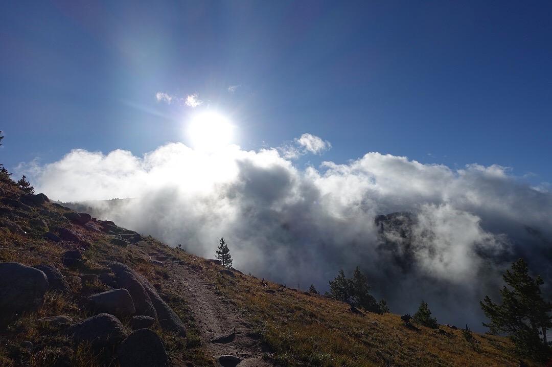 A sunlit landscape showing a winding dirt path along a hillside, surrounded by rocks and patches of grass. Above, wispy clouds float against a bright blue sky with the sun shining brightly, creating a serene and picturesque scene. Monarch Crest Trail mountain bike trail.