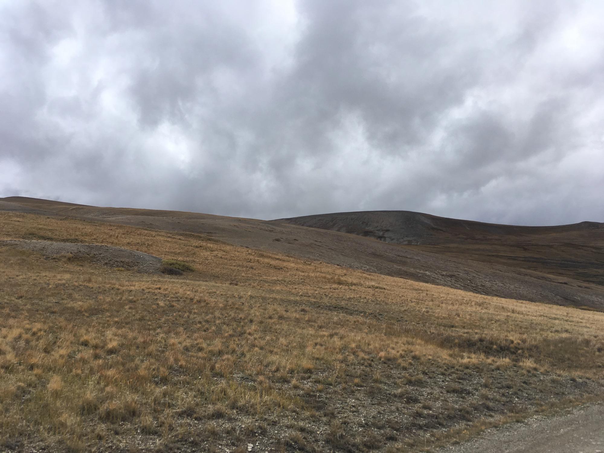 A scenic view of a grassy landscape under a cloudy sky, featuring rolling hills and a mixture of dry and rocky terrain. The atmosphere appears overcast, creating a dramatic contrast between the soft grass and the rugged hills in the background. Weston Pass Road / Co Rd #22 mountain bike trail.