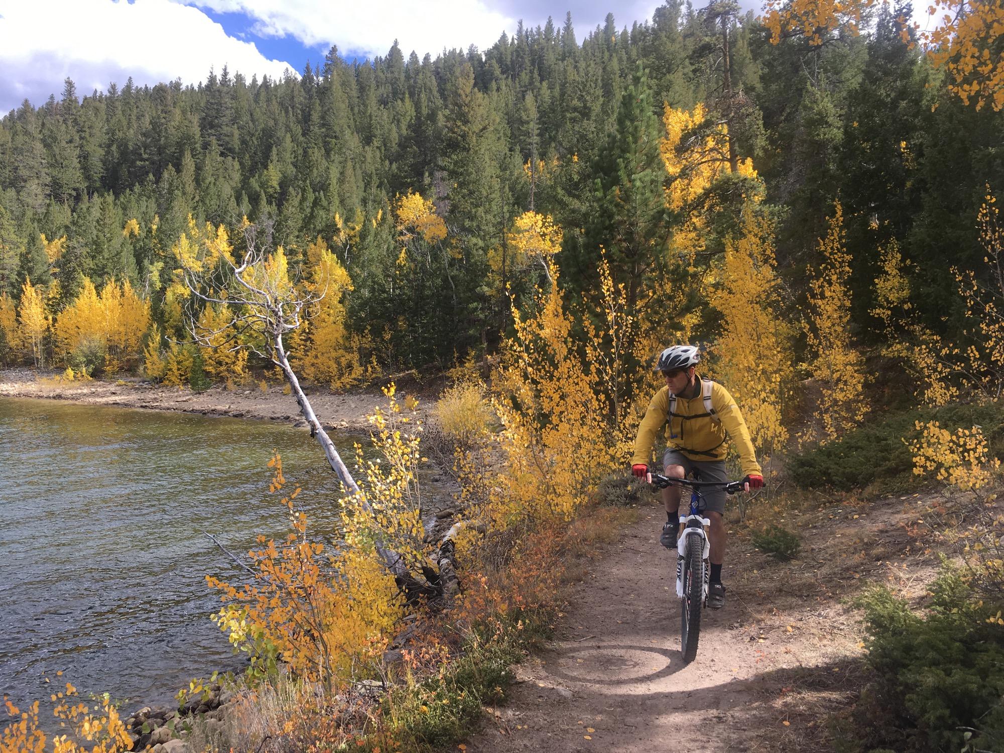 A person riding a mountain bike along a dirt trail beside a lake, surrounded by vibrant yellow and green trees in an autumn landscape. The sky is partly cloudy, adding to the scenic outdoor atmosphere. Twin Lakes Loop mountain bike trail.