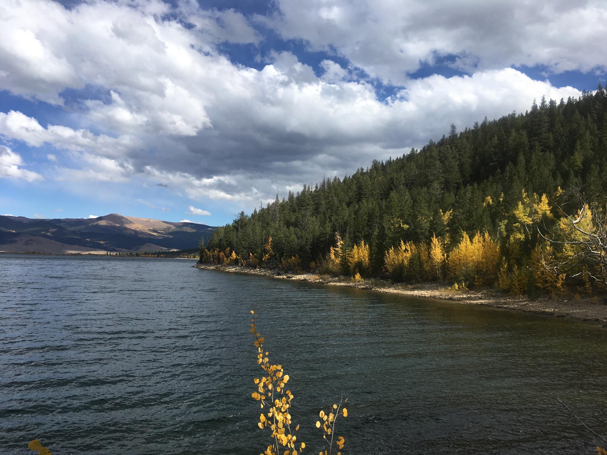 A serene view of a lake surrounded by a hillside of evergreen trees, with patches of golden foliage visible along the shore. The sky is partly cloudy, featuring a mix of blue and white clouds, and distant mountains are visible in the background. Twin Lakes Loop mountain bike trail.