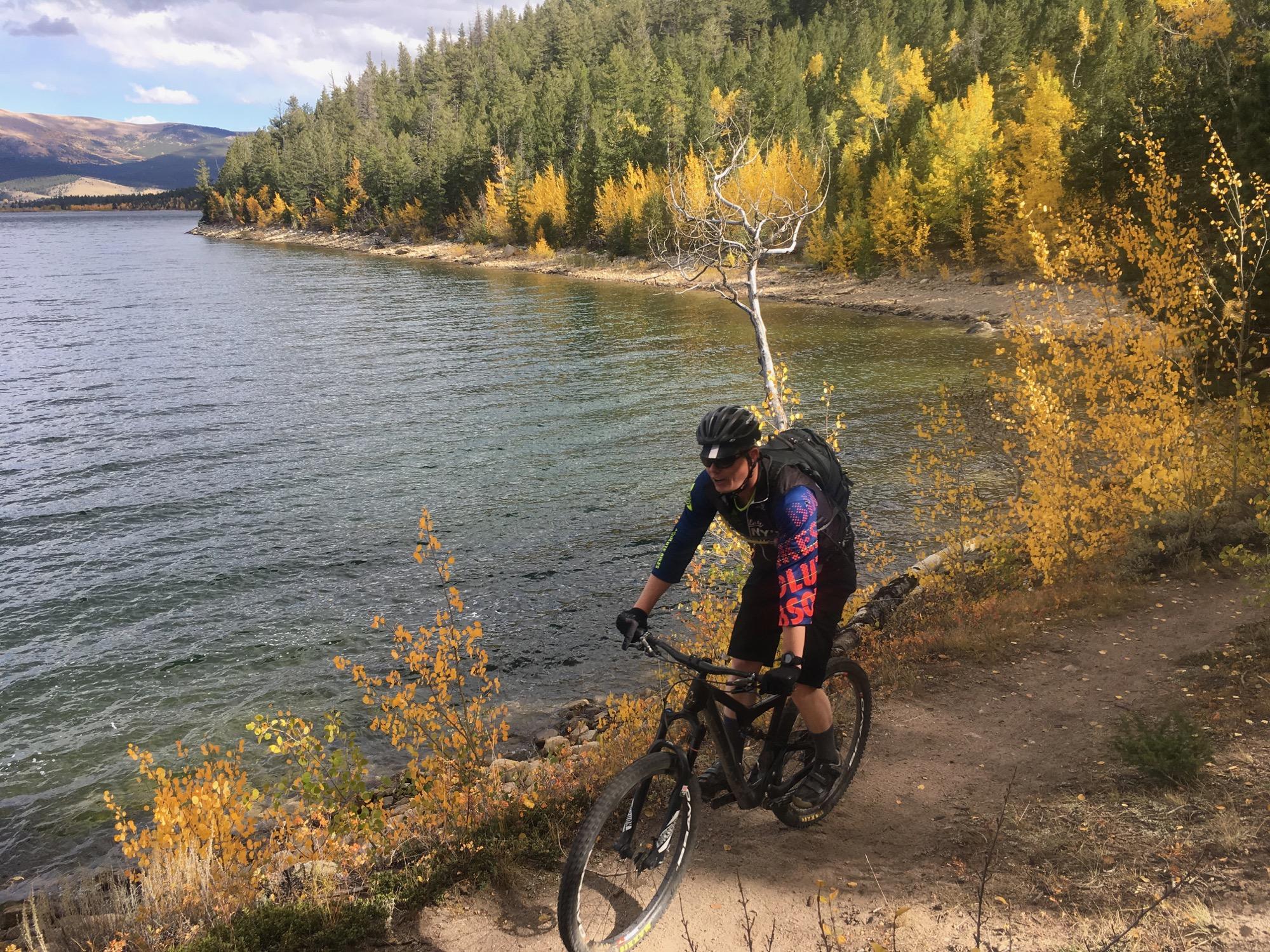 A mountain biker navigates a dirt path alongside a scenic lake, surrounded by lush green pine trees and vibrant autumn foliage. The water reflects the clear sky and distant mountains, creating a picturesque outdoor setting perfect for biking adventures. Colorado Trail: Clear Creek Thd to Lake View CG / Hwy 82 mountain bike trail.