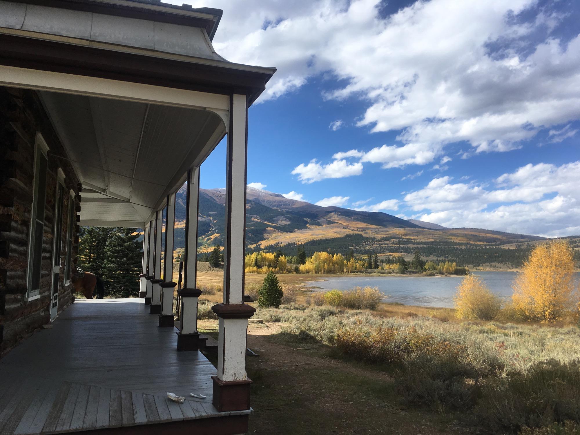 A scenic view from a rustic porch overlooking a lake surrounded by autumn foliage, with mountains in the background under a partly cloudy sky. Twin Lakes Loop mountain bike trail.