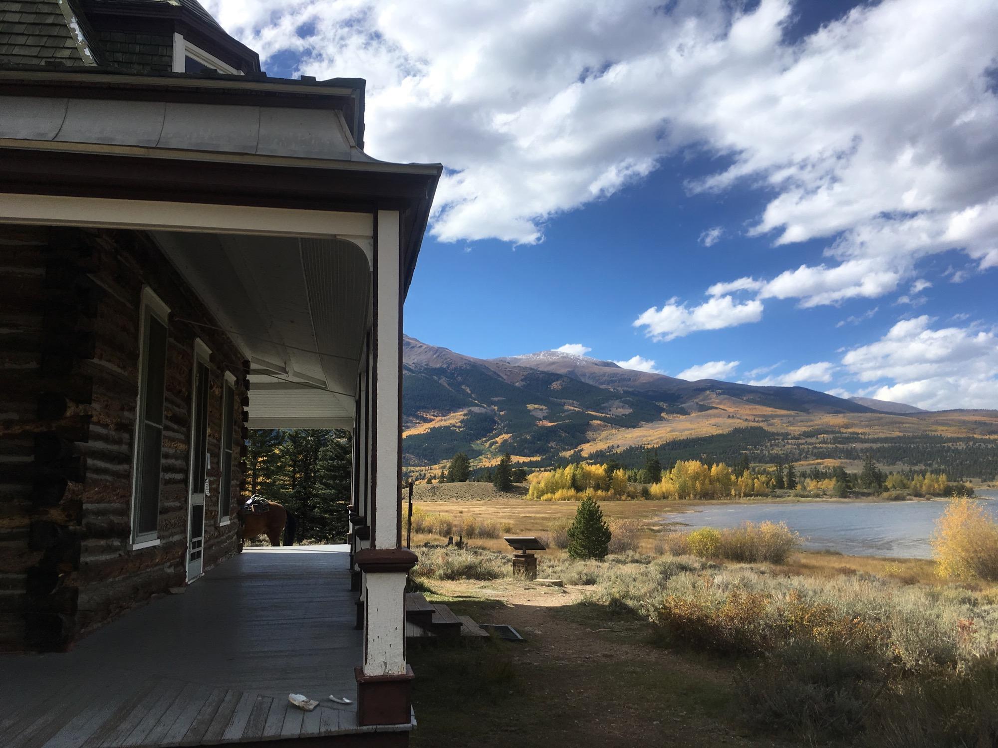 A scenic view from a porch featuring a log cabin, with mountains and a lake in the background. The landscape is adorned with autumn foliage in shades of yellow and green under a partly cloudy blue sky. A horse is visible near the edge of the porch. Twin Lakes Loop mountain bike trail.
