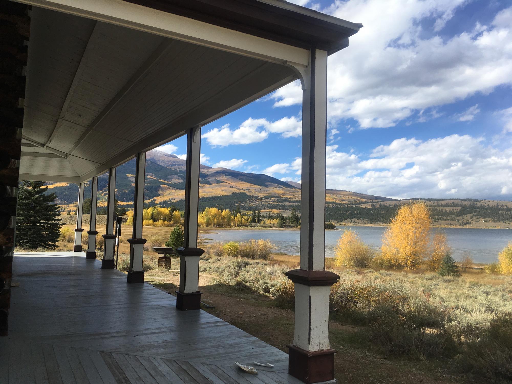 View from a porch overlooking a tranquil lake surrounded by autumn foliage and mountains under a partly cloudy sky. The scene features a wooden railing and columns of the porch in the foreground, with vibrant yellow and green trees lining the shore and distant hills. Colorado Trail: Clear Creek Thd to Lake View CG / Hwy 82 mountain bike trail.