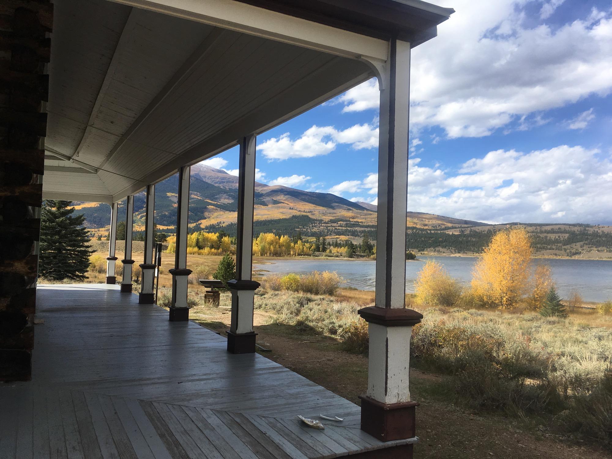 A scenic view from a porch overlooking a calm river, surrounded by mountains and vibrant autumn foliage. The sky features scattered clouds, and the landscape includes trees with golden leaves, adding a warm touch to the tranquil setting. Colorado Trail: Clear Creek Thd to Lake View CG / Hwy 82 mountain bike trail.