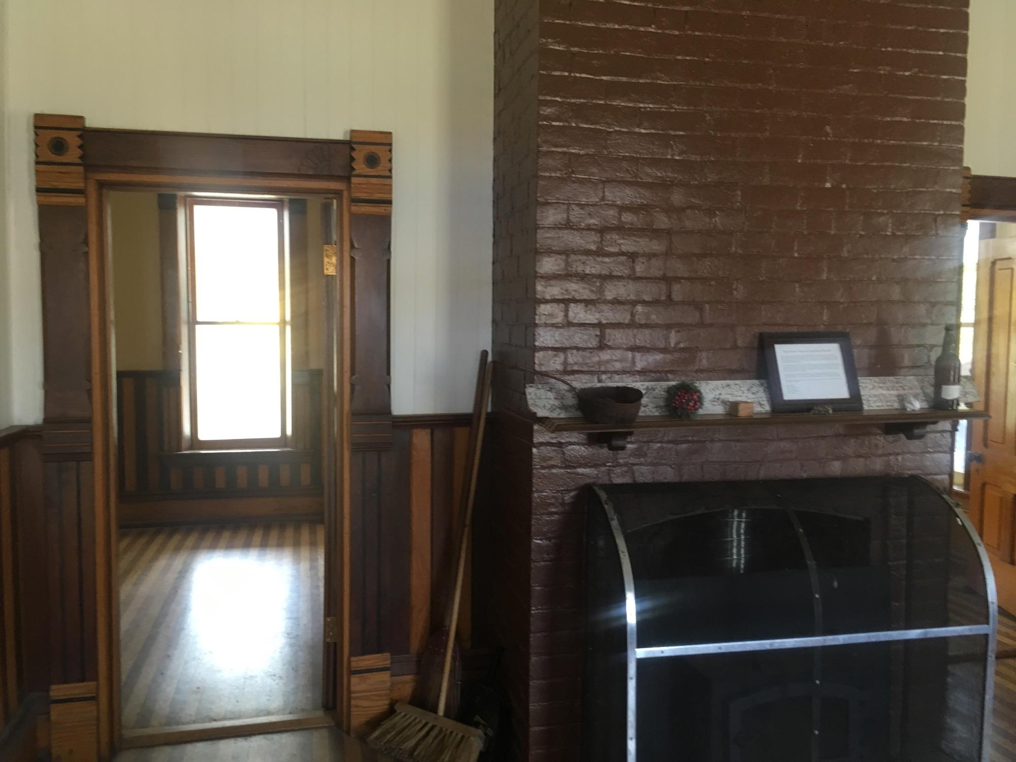 A cozy interior space featuring a wooden doorway with decorative trim leading to a room with natural light from a window. The adjacent wall displays a brown brick fireplace with a wooden mantel adorned with a framed document, decorative items, and a bottle. The floor is visible with wooden planks, contributing to the warm ambiance of the room. Colorado Trail: Clear Creek Thd to Lake View CG / Hwy 82 mountain bike trail.