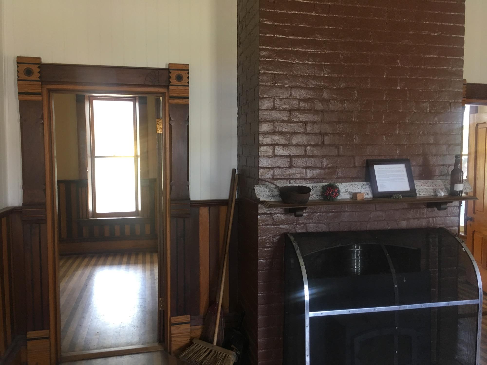 Interior view of a room featuring a wooden door frame with decorative details, leading to a brightly lit space with a window. In the foreground, there is a brick fireplace with a wooden mantel displaying a framed document, a small decorative item, and a bottle. The room has wooden flooring and paneling, contributing to a cozy atmosphere. Twin Lakes Loop mountain bike trail.