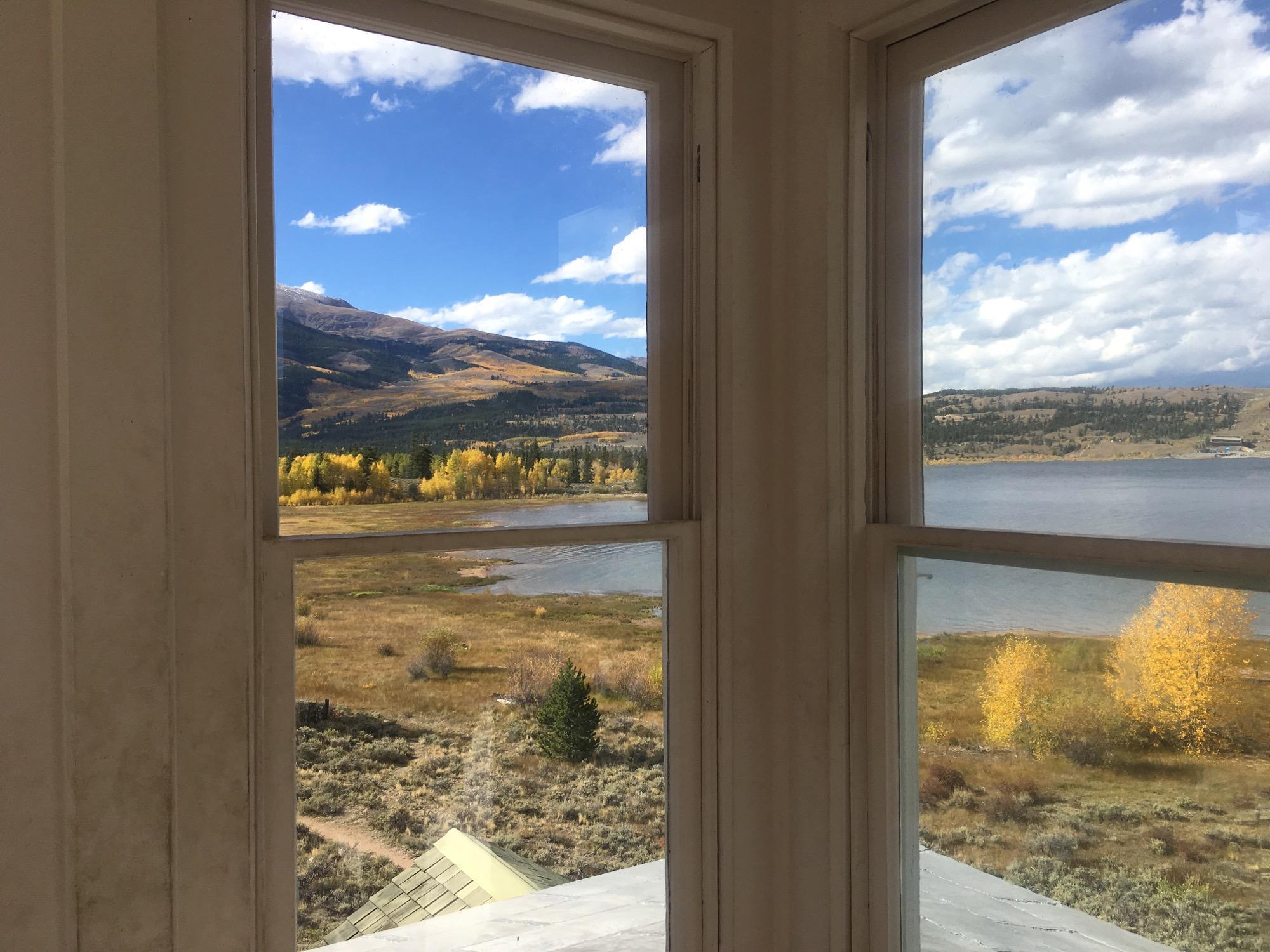 A scenic view from a window, showcasing mountains in the background with rugged landscapes and colorful autumn foliage in shades of orange and yellow. The water of a lake is visible below, framed by the window panes. The sky is partly cloudy, adding to the serene atmosphere of the scene. Twin Lakes Loop mountain bike trail.