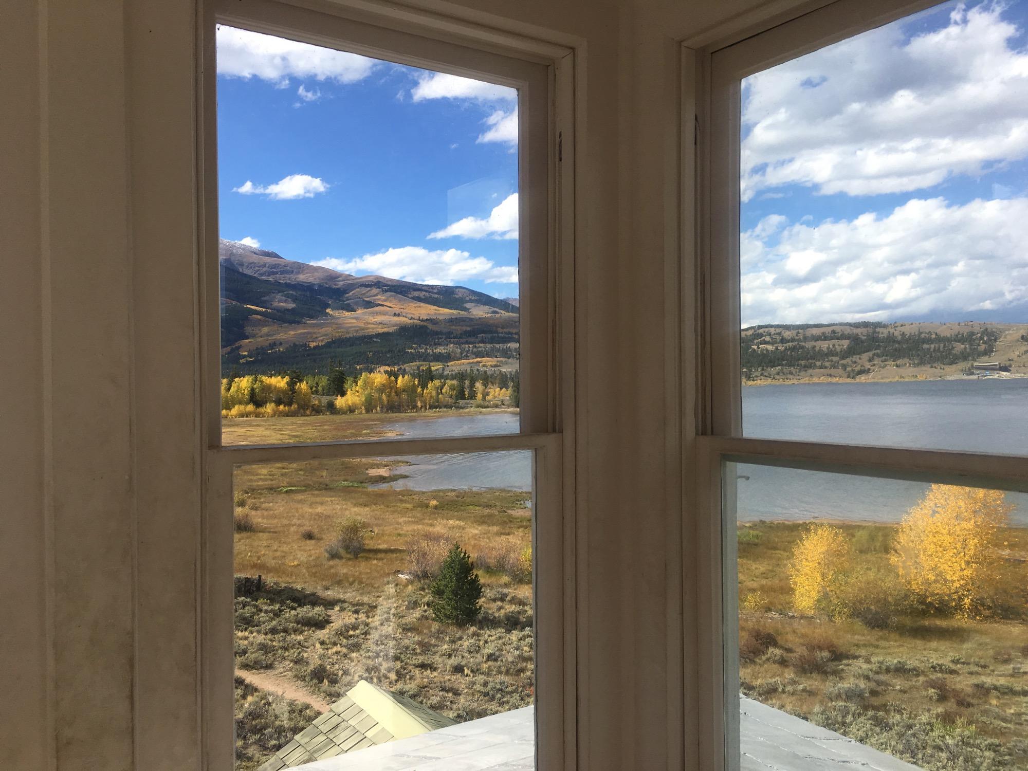 View through window panes showcasing a scenic landscape of mountains, a serene lake, and autumn foliage. Bright blue skies with scattered clouds enhance the picturesque setting, featuring trees with golden leaves along the shoreline and rolling hills in the background. Twin Lakes Loop mountain bike trail.