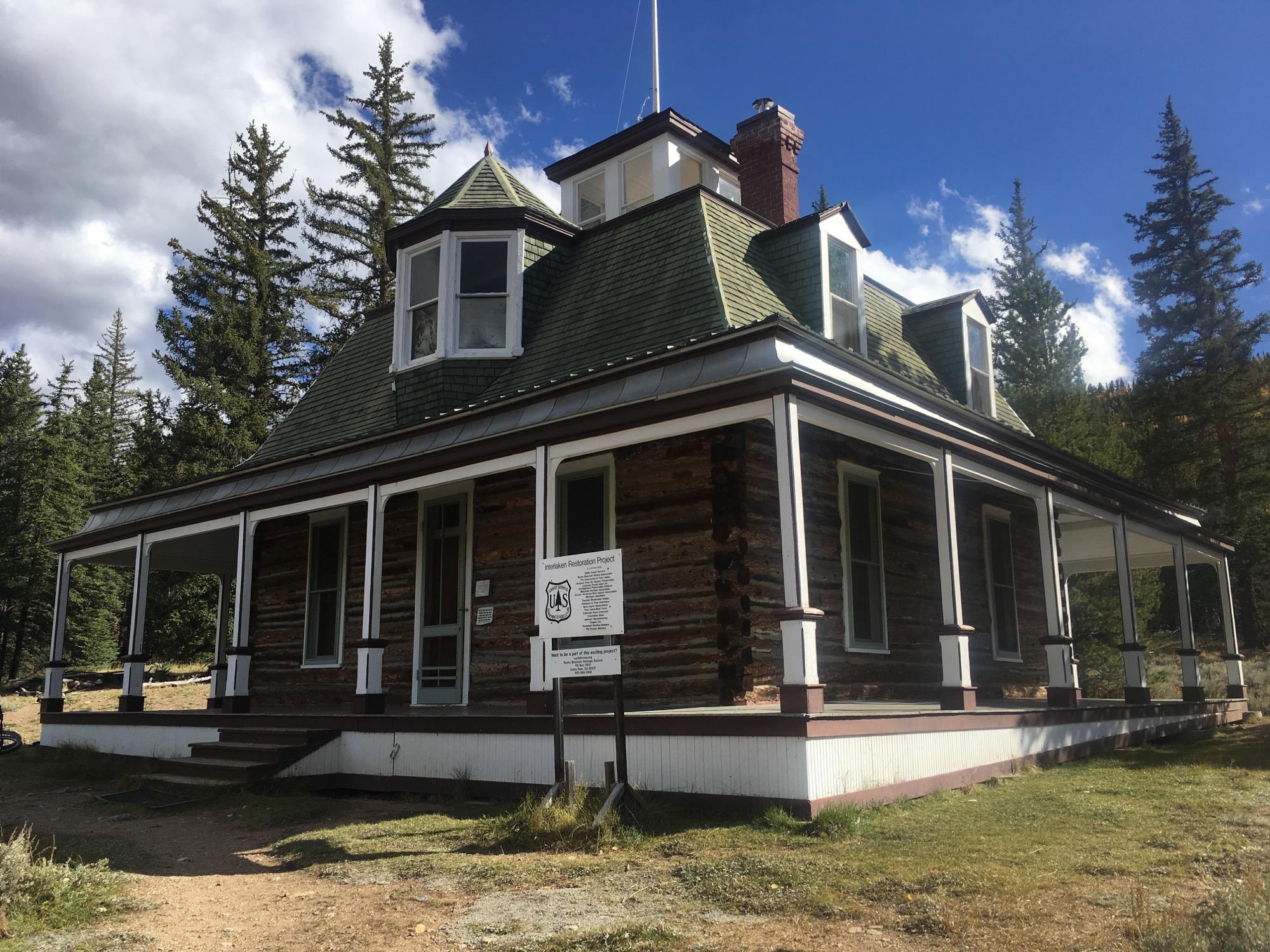 A historic log cabin with a green gabled roof, featuring multiple windows and a wraparound porch. The building is surrounded by tall pine trees and grassy areas, set against a backdrop of partly cloudy blue sky. A sign indicating a restoration project is visible near the entrance. Twin Lakes Loop mountain bike trail.