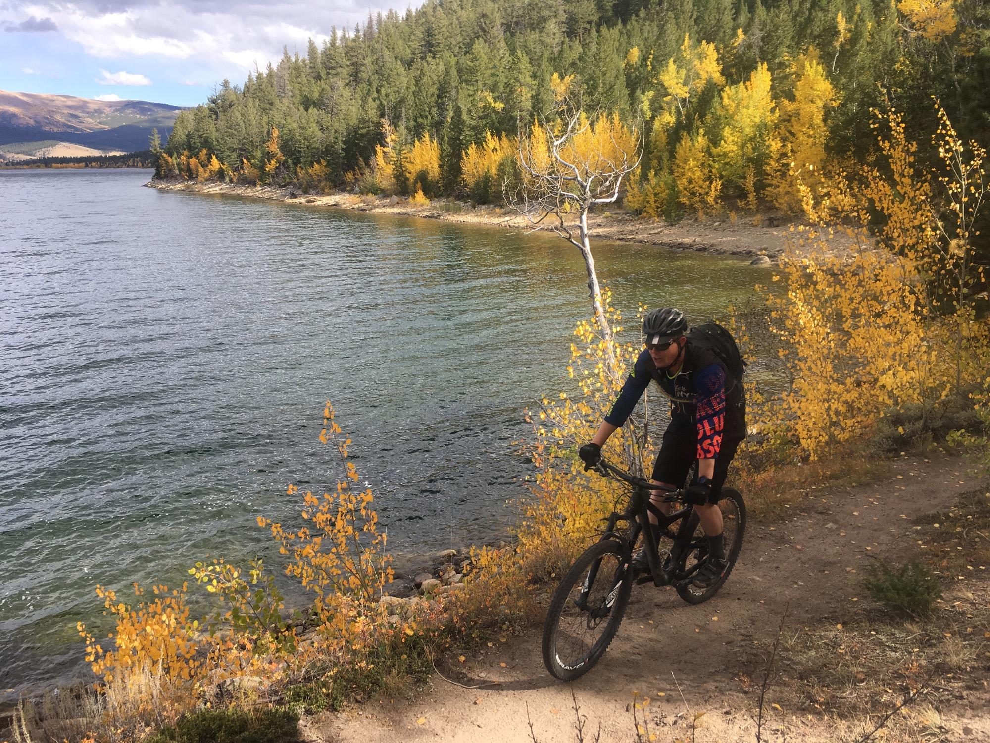 A mountain biker navigates a dirt trail along the edge of a lake surrounded by vibrant autumn foliage. The scene features a mix of lush green and bright yellow trees, with a clear blue sky and distant mountains in the background. Twin Lakes Loop mountain bike trail.